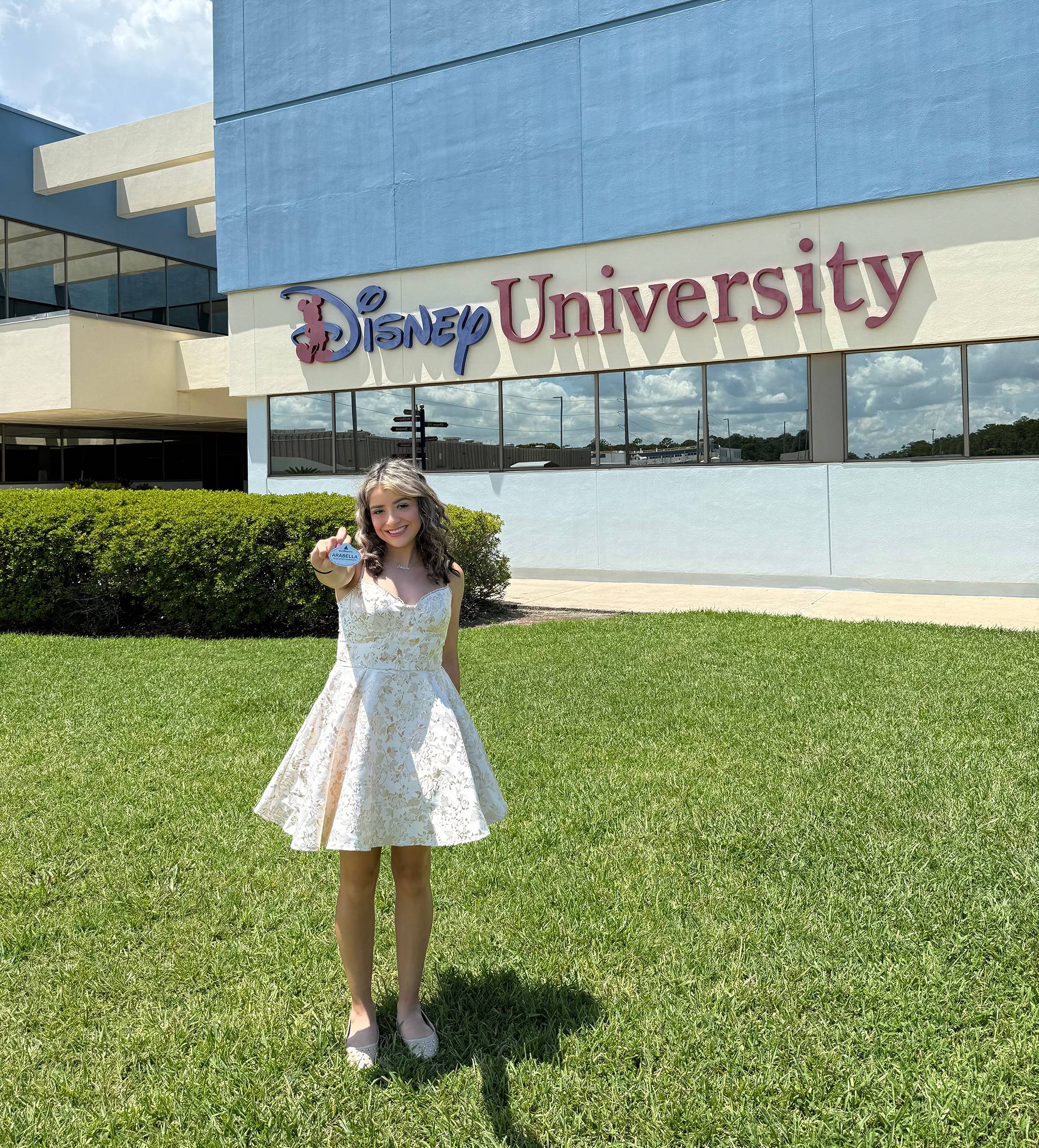 woman in white dress stands outside the disney university holding her name tag out towards the camera