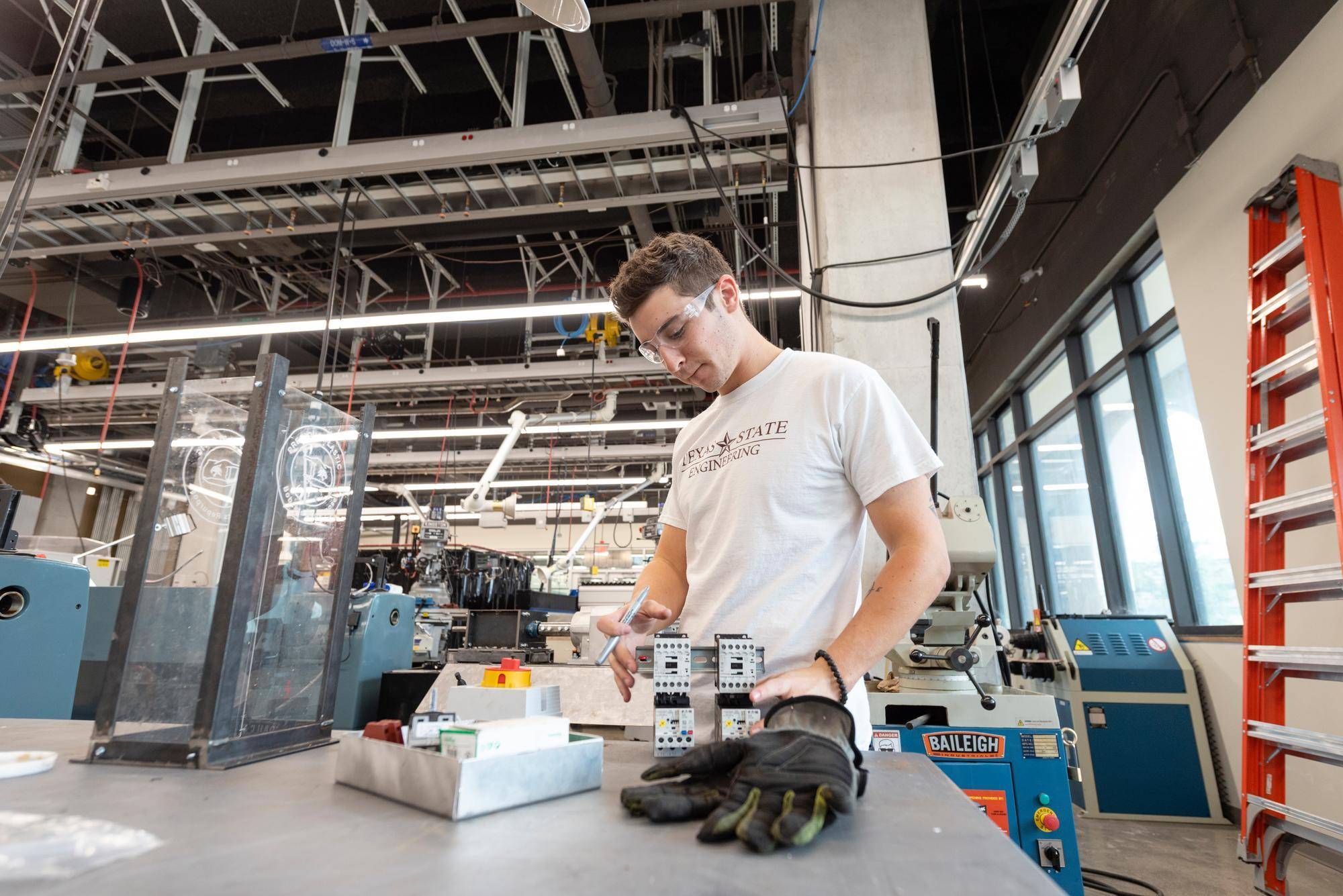 a student wearing safety goggles and a white texas state t-shirt works on a machine in a workshop