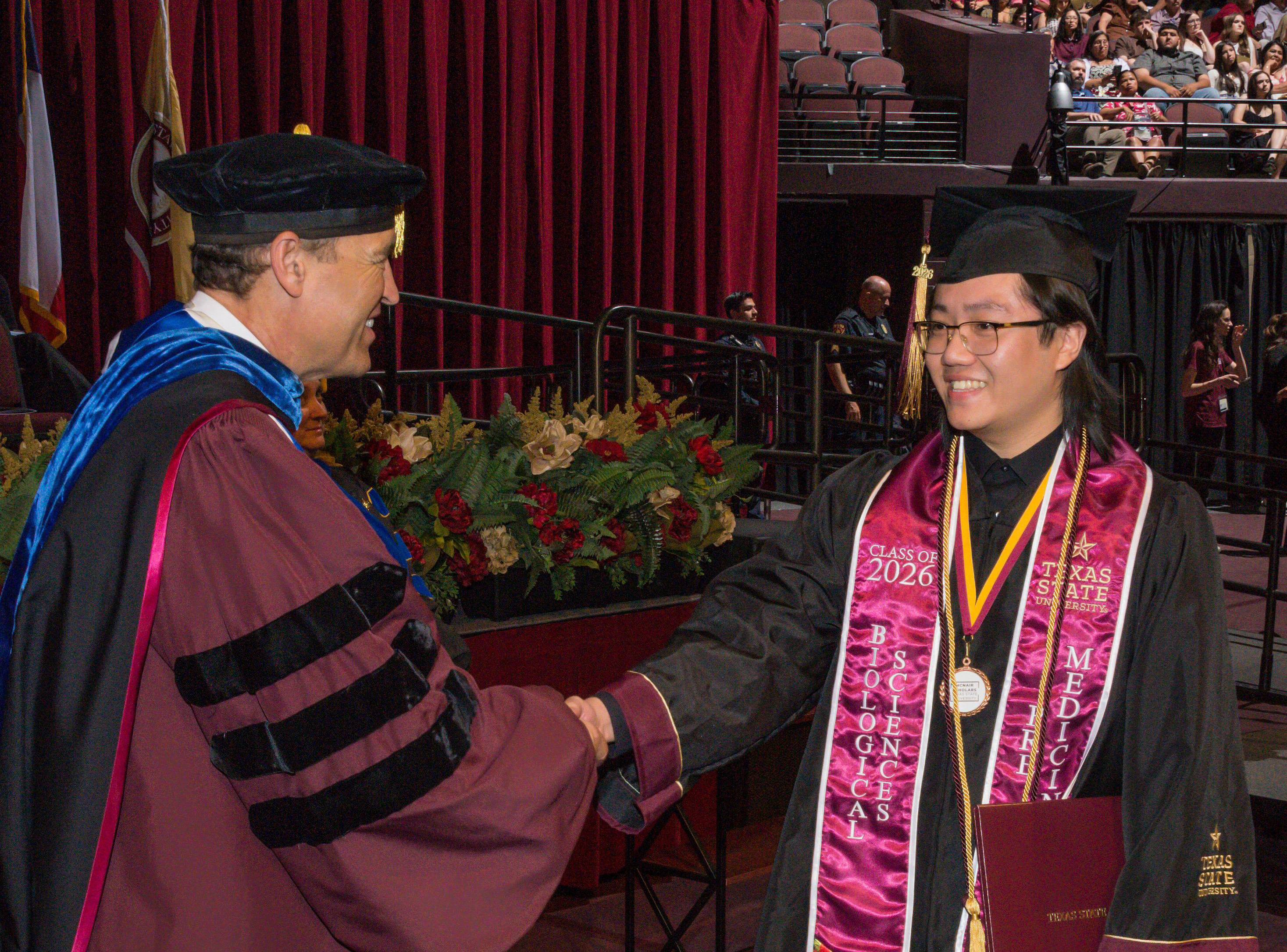 man in graduation regalia shakes hand of university president during commencement ceremony