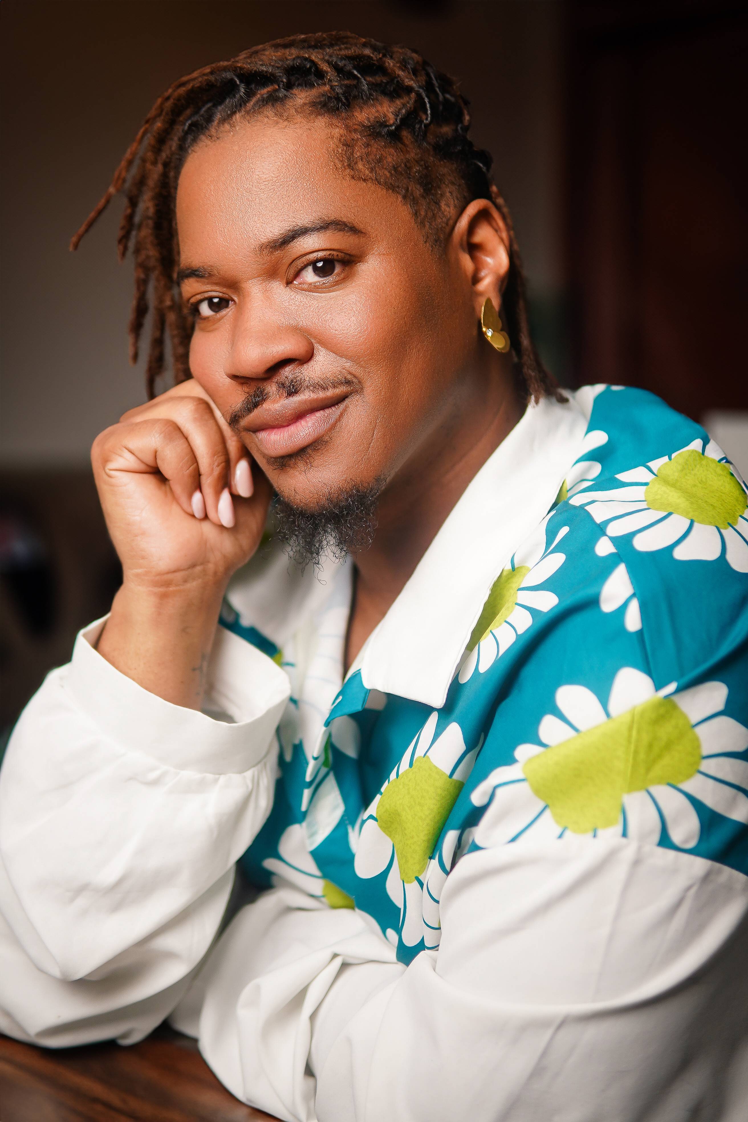 Headshot of Deonté Warren smiling warmly with his hand resting on his chin, wearing a white button-up shirt with a green and yellow floral pattern.
