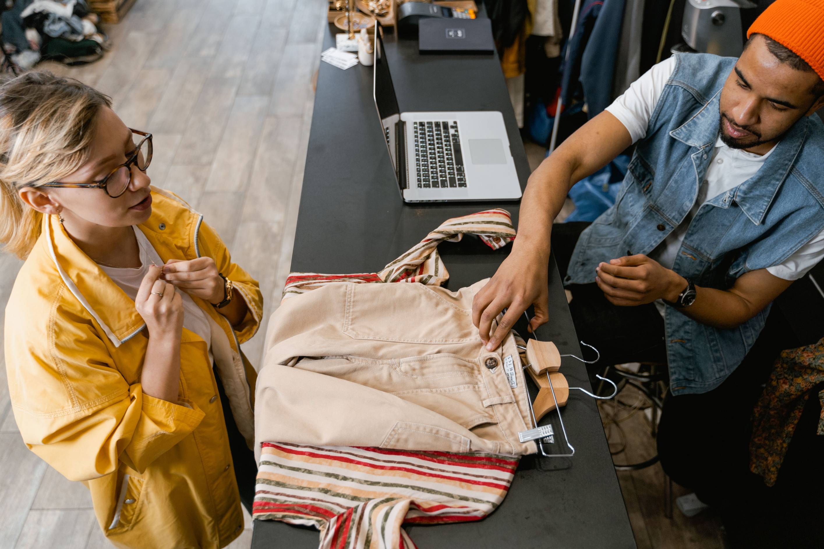 Overhead view of a customer and retail employee discussing a clothing purchase at a checkout counter. A pair of beige pants is displayed on the counter beside a striped tote bag and laptop.