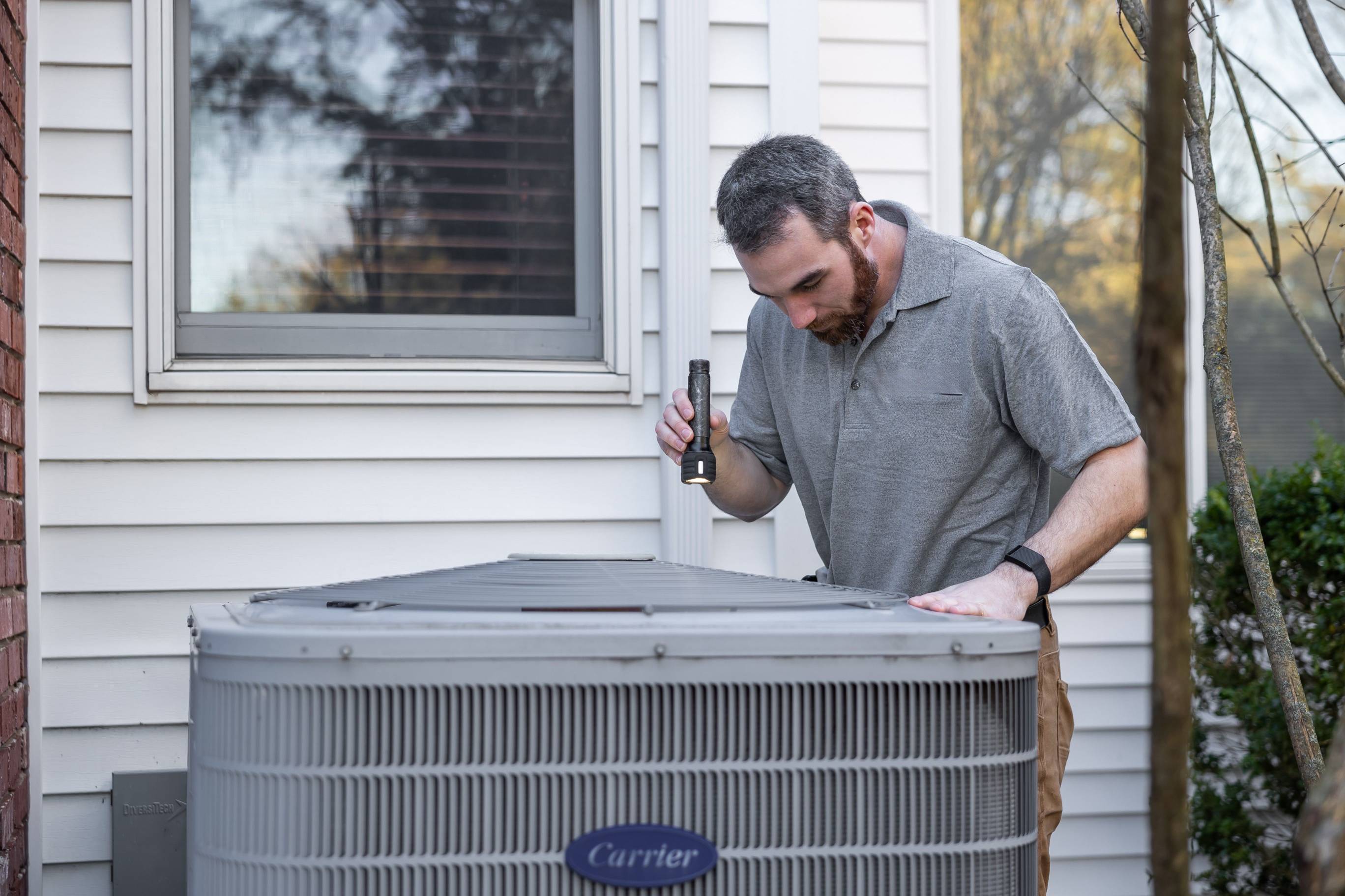 HVAC technician inspecting an outdoor air conditioning unit beside a home, using a flashlight to examine the equipment. The technician stands next to a large residential cooling system, representing skilled trades, home maintenance, HVAC services, repair work, and technical field careers.