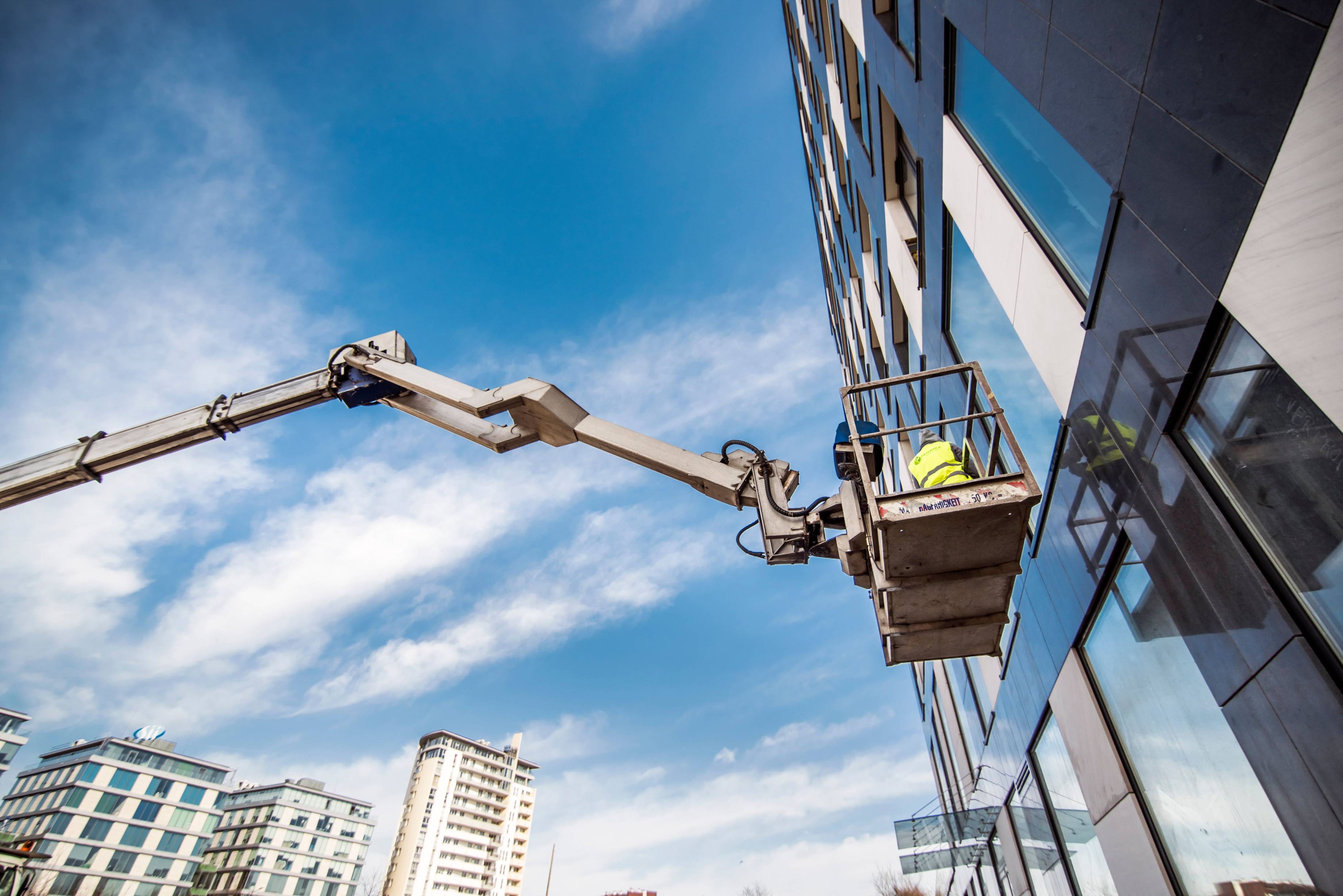Worker in a high-reach lift performs maintenance on the exterior of a modern multi-story building. Elevated beside large windows, the worker uses specialized equipment to access upper levels. The image represents building maintenance, property management, commercial services, safety practices, and skilled trades work.