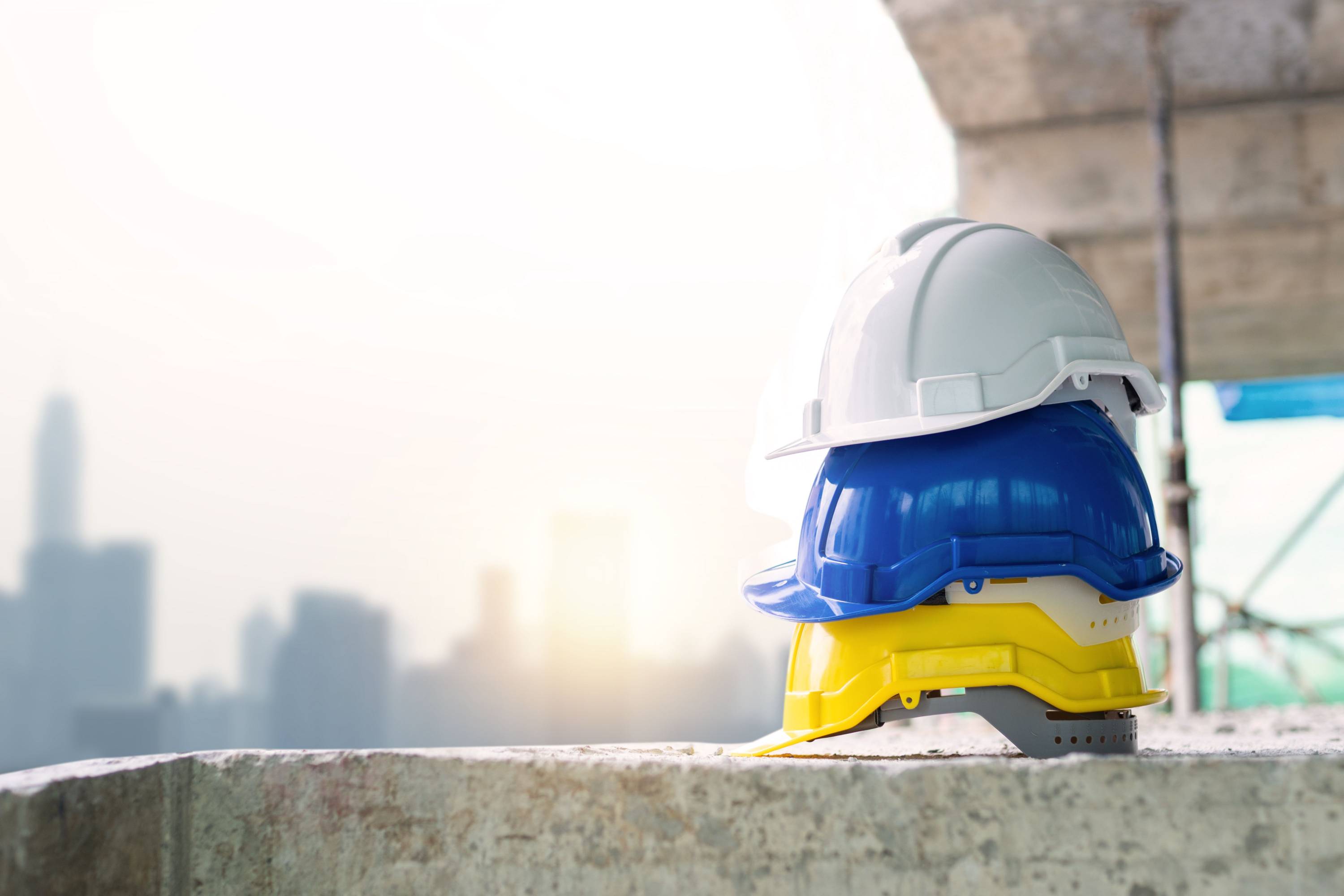 Stack of white, blue, yellow, and gray hard hats resting on a construction site ledge with a city skyline in the background. The image symbolizes workplace safety, construction, skilled trades, infrastructure projects, and careers in the building industry, emphasizing the importance of protective equipment on the job.