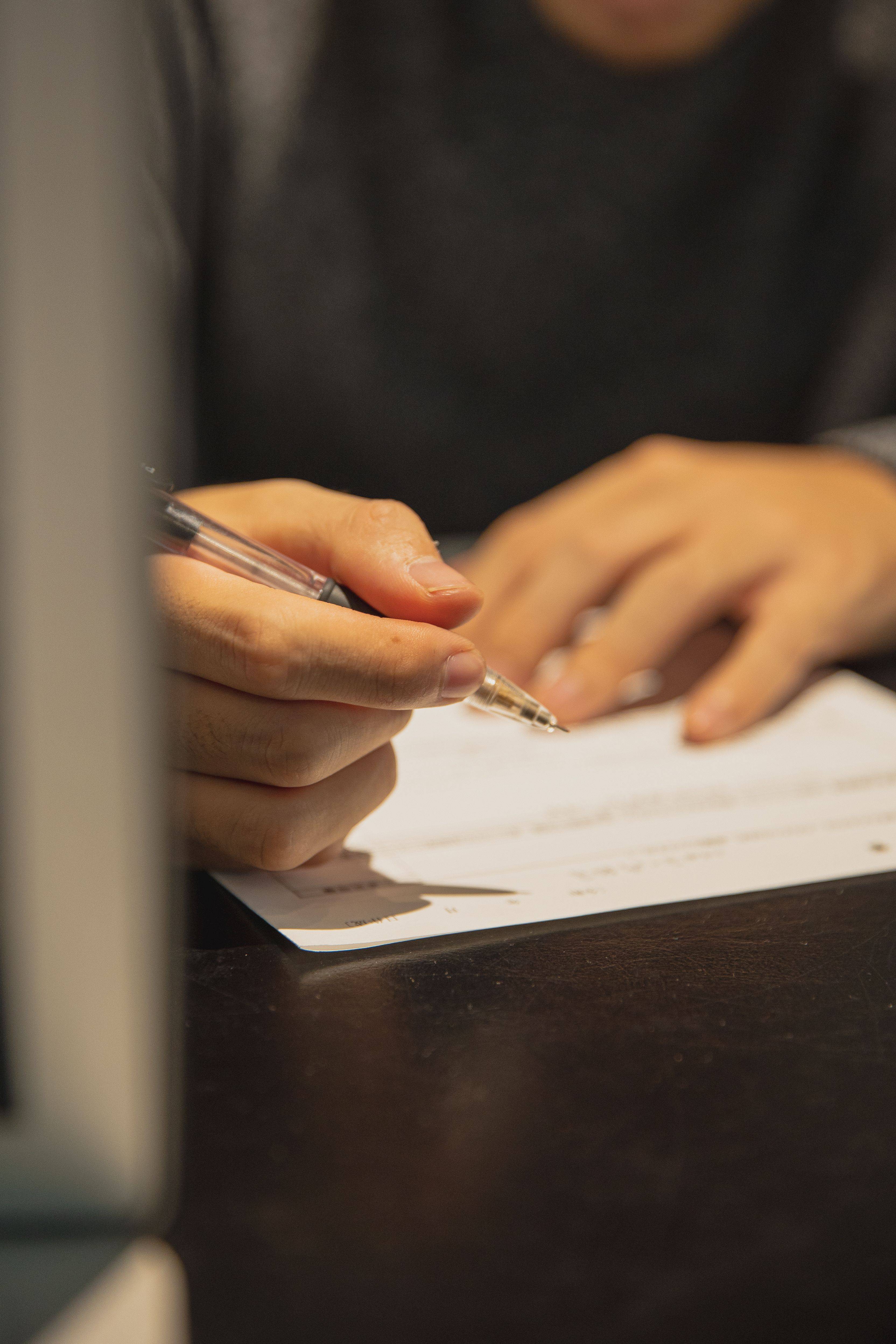 Close-up of a person writing on a document with a pen at a desk. The focus is on the hand and paperwork, with the background softly blurred. The image represents completing forms, business planning, recordkeeping, contracts, applications, or administrative tasks in a professional setting.