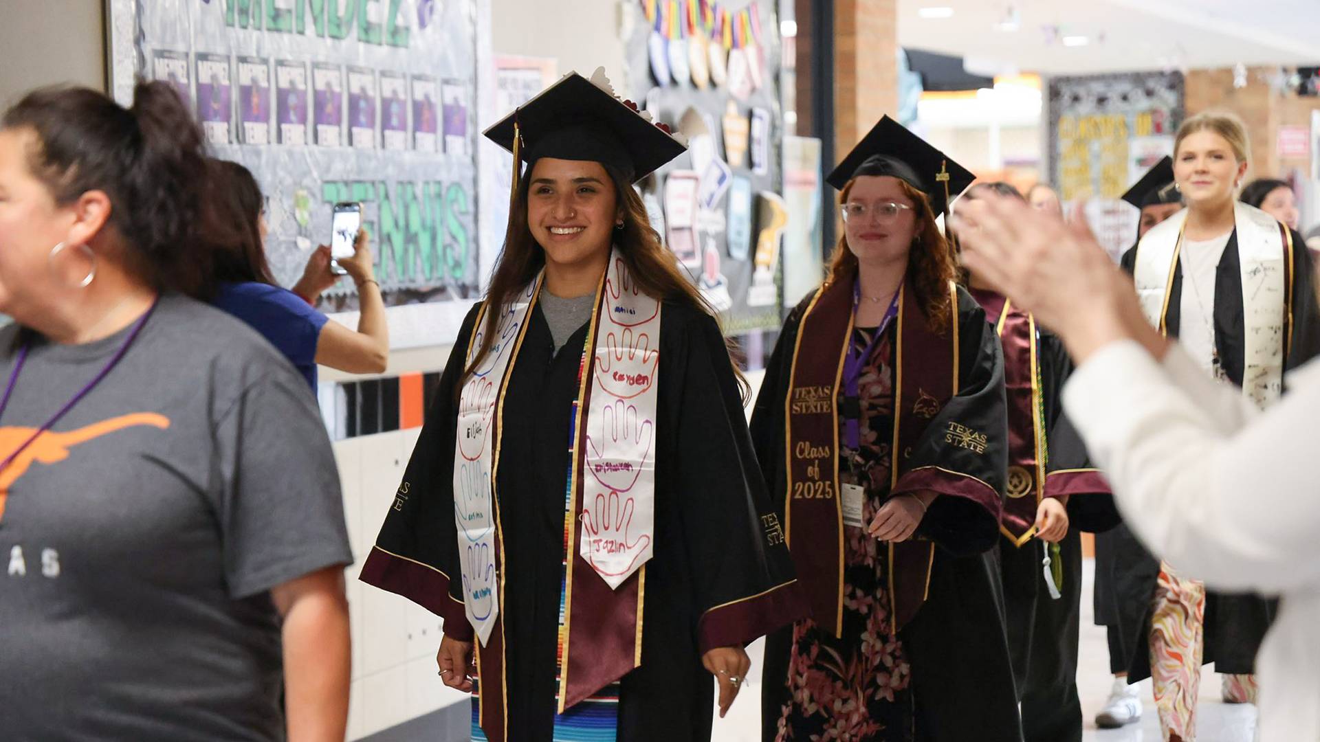 two students in graduation regalia walking down hallway of elementary school