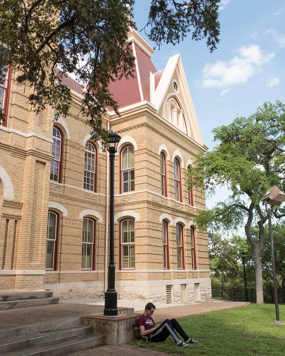 Student leaning against a lamppost, reading a book, outside of the Old Main building on the San Marcos campus 
