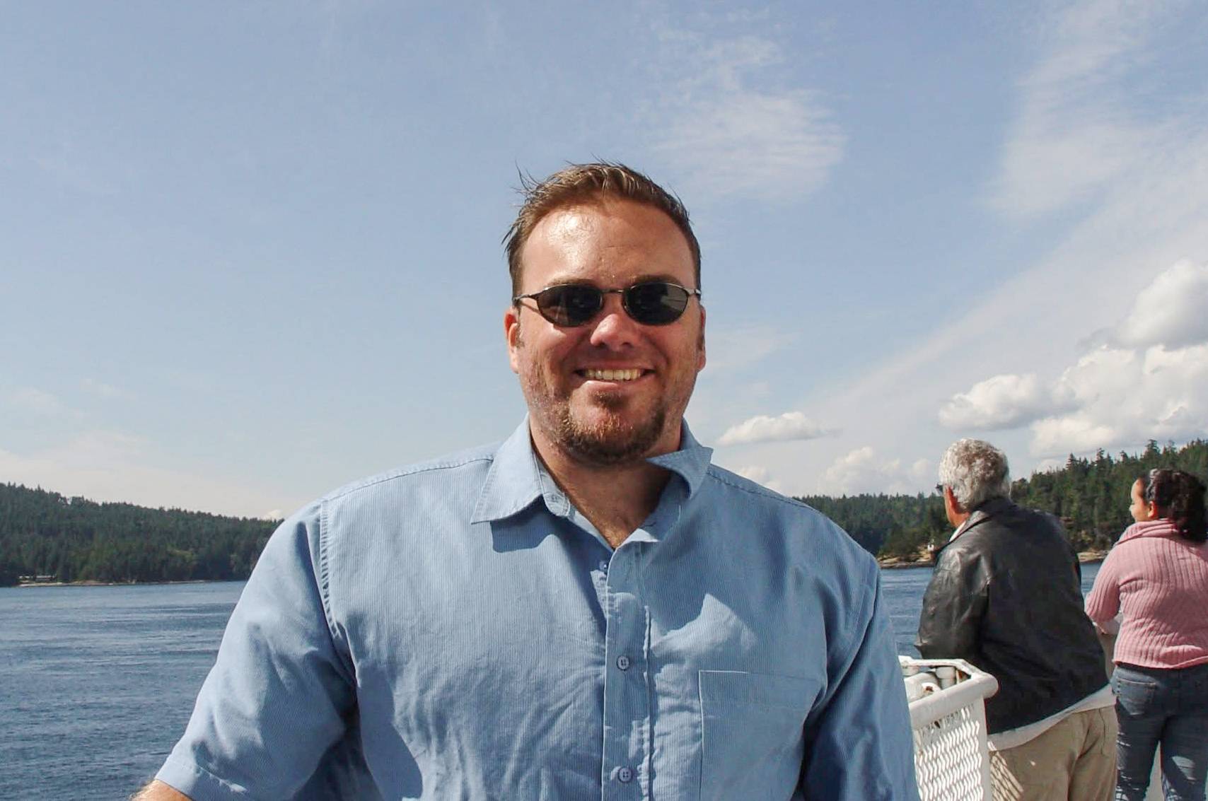 Brad Monk, wearing sunglasses and a blue button up shirt, stands outside in front of a body of water.