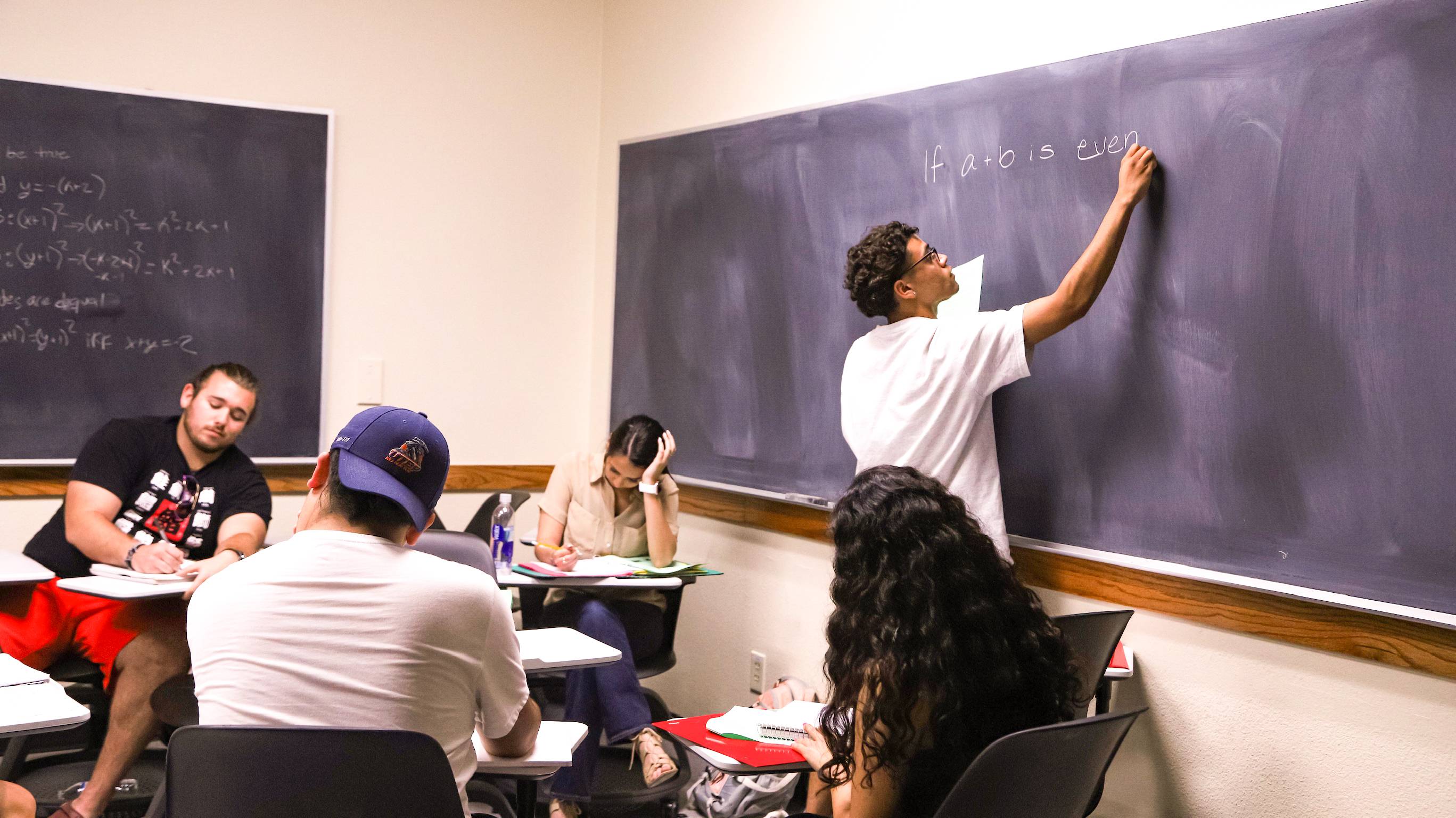 Students sit in a classroom while one rights an equation on a black board. 
