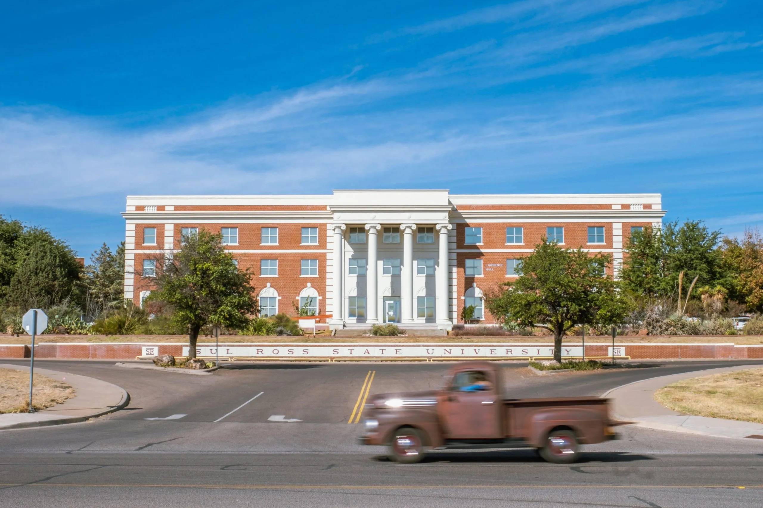 a truck drives by a orange brick building with white pillars