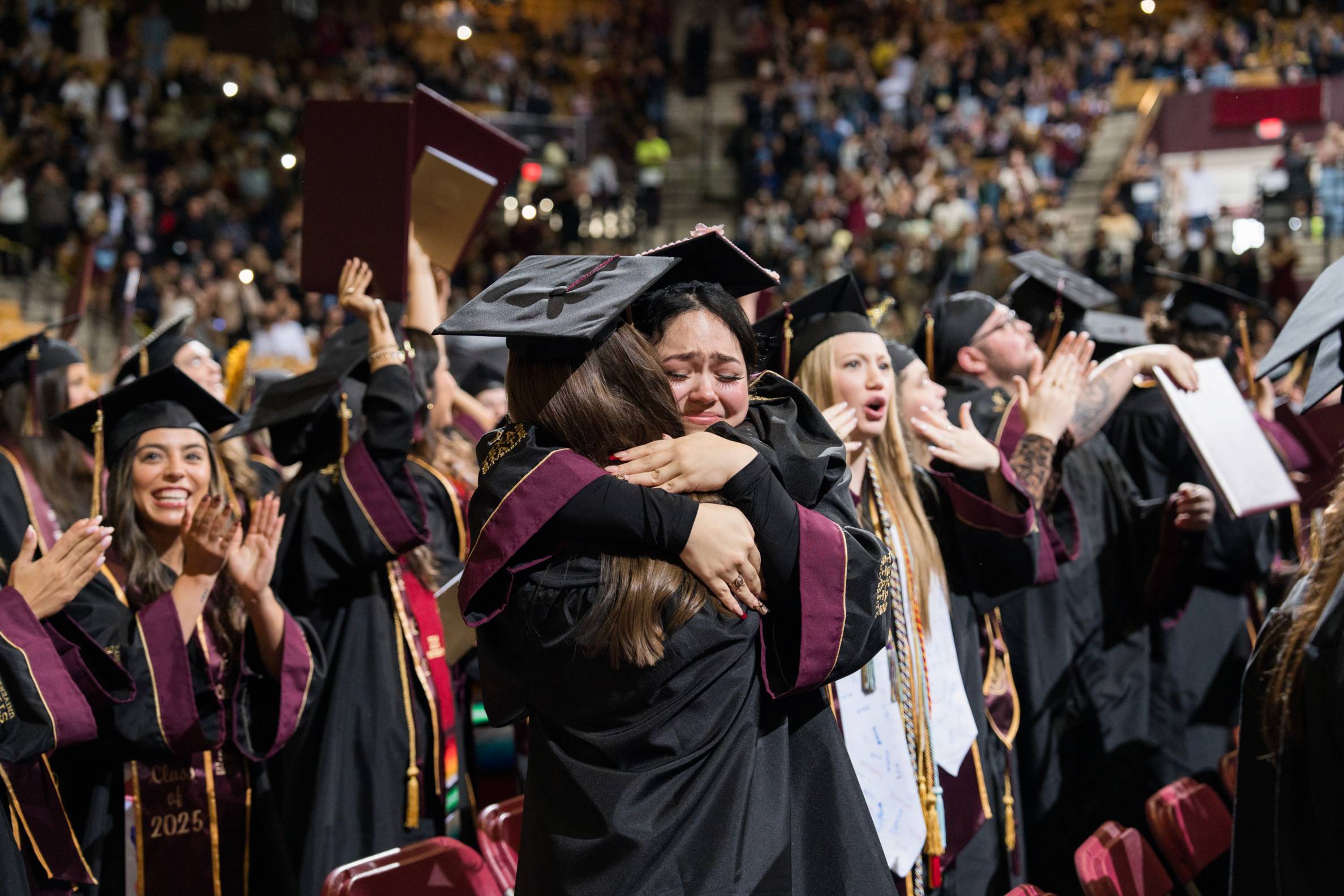 Two students hug while wearing caps and gowns at commencement surrounded by fellow graduates who are cheering. 
