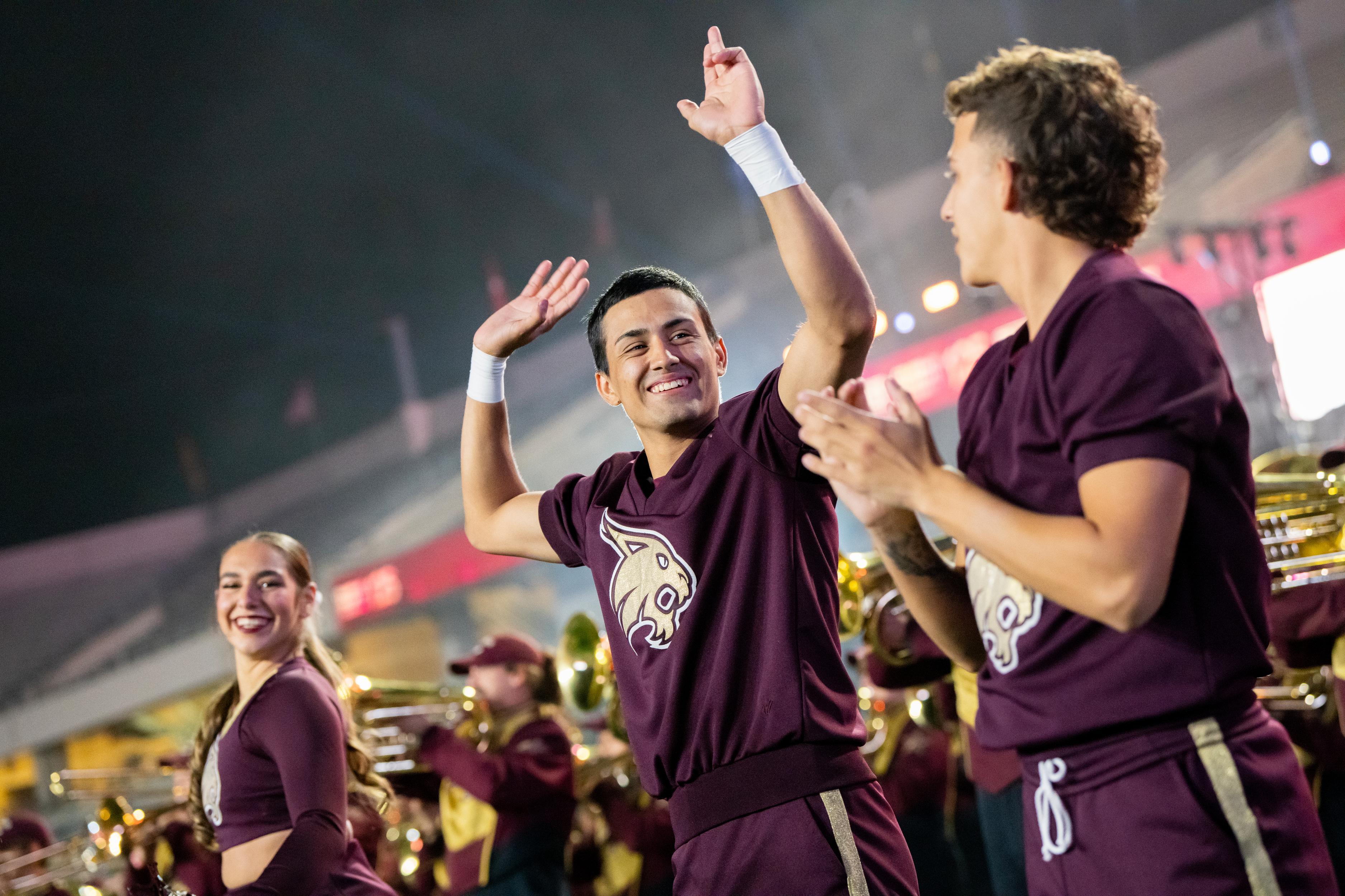 male cheerleader holding up txst hand sizes above his head as he looks to another cheerleader and smiles