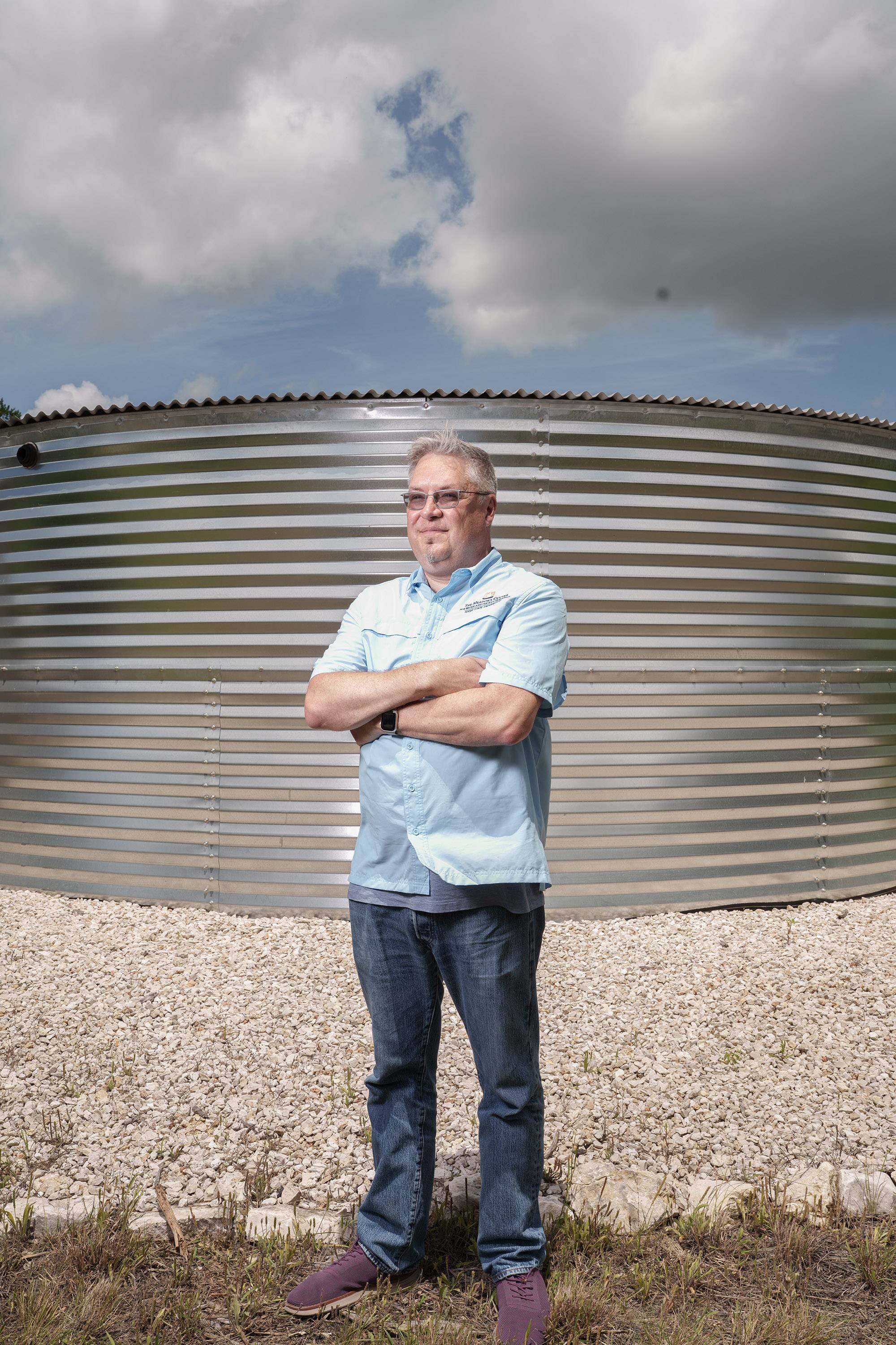 Dr. Robert Mace stands with arms crossed in front of a large water storage tank made from corrugated metal.