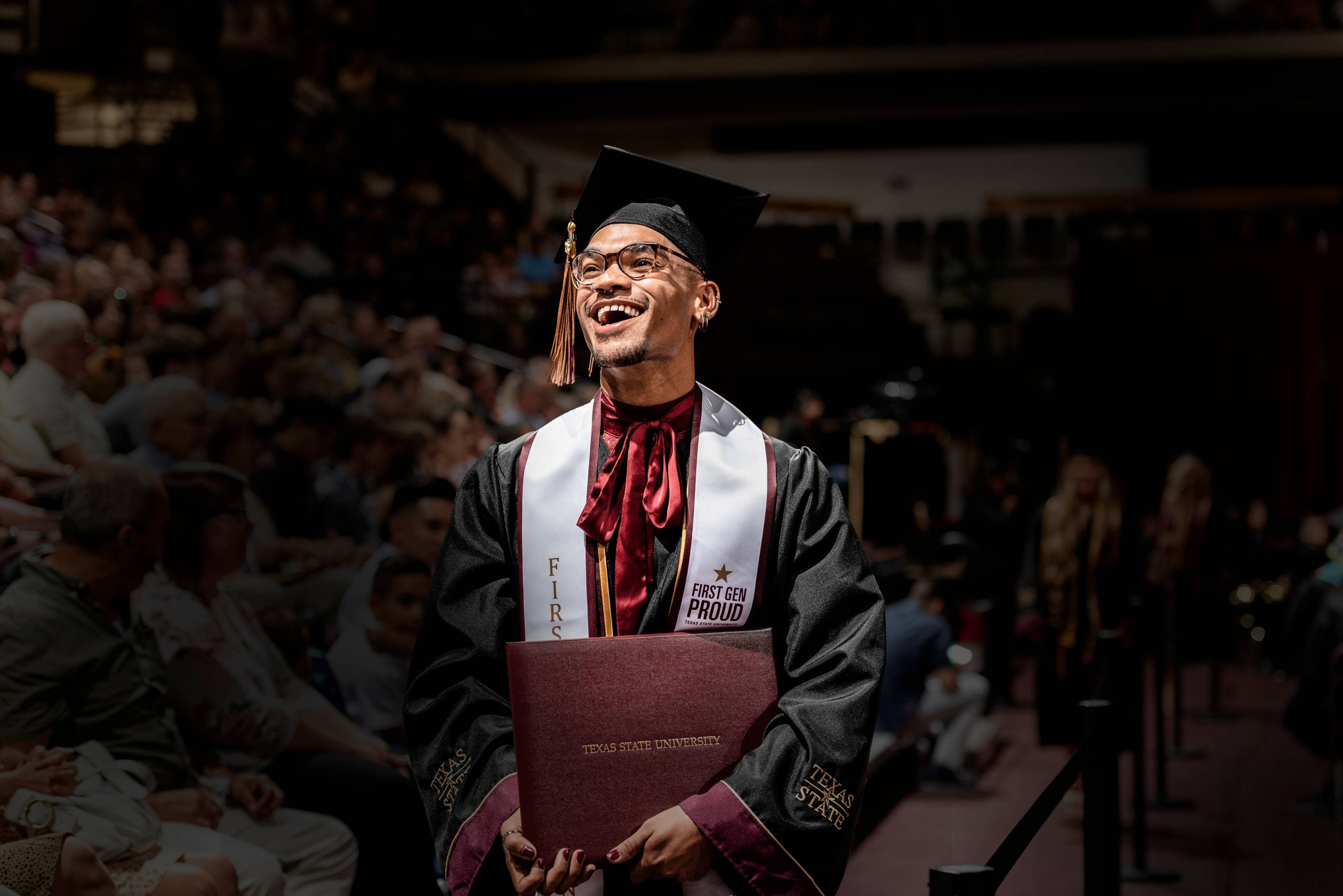 A student in cap and gown smiles up at the crowd at commencement. He is wearing a stole that reads "first generation."
