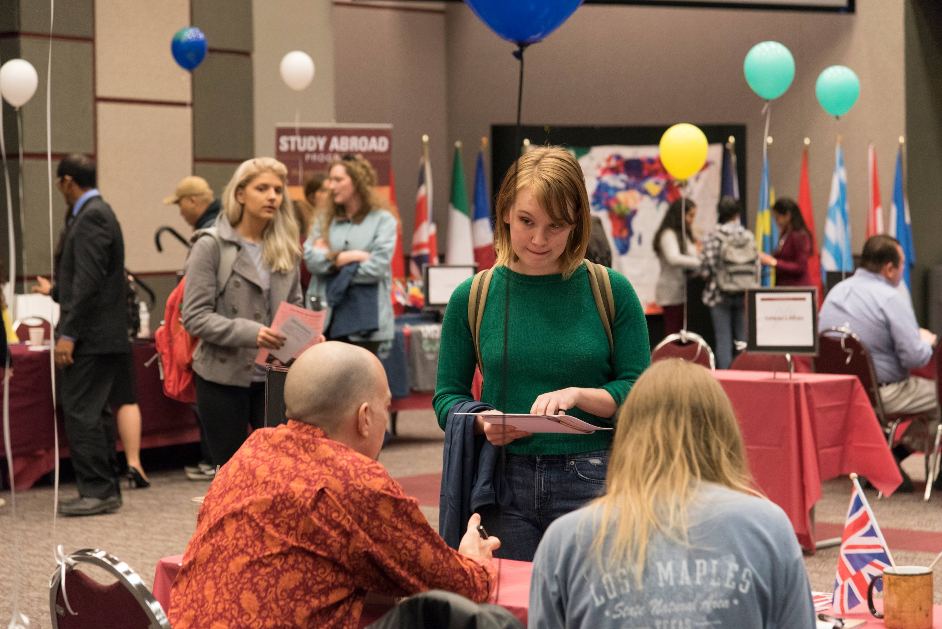 A student holds papers and talks with two people seated at a table. There are international flags, maps, and balloons decorating the room of a study abroad fair.