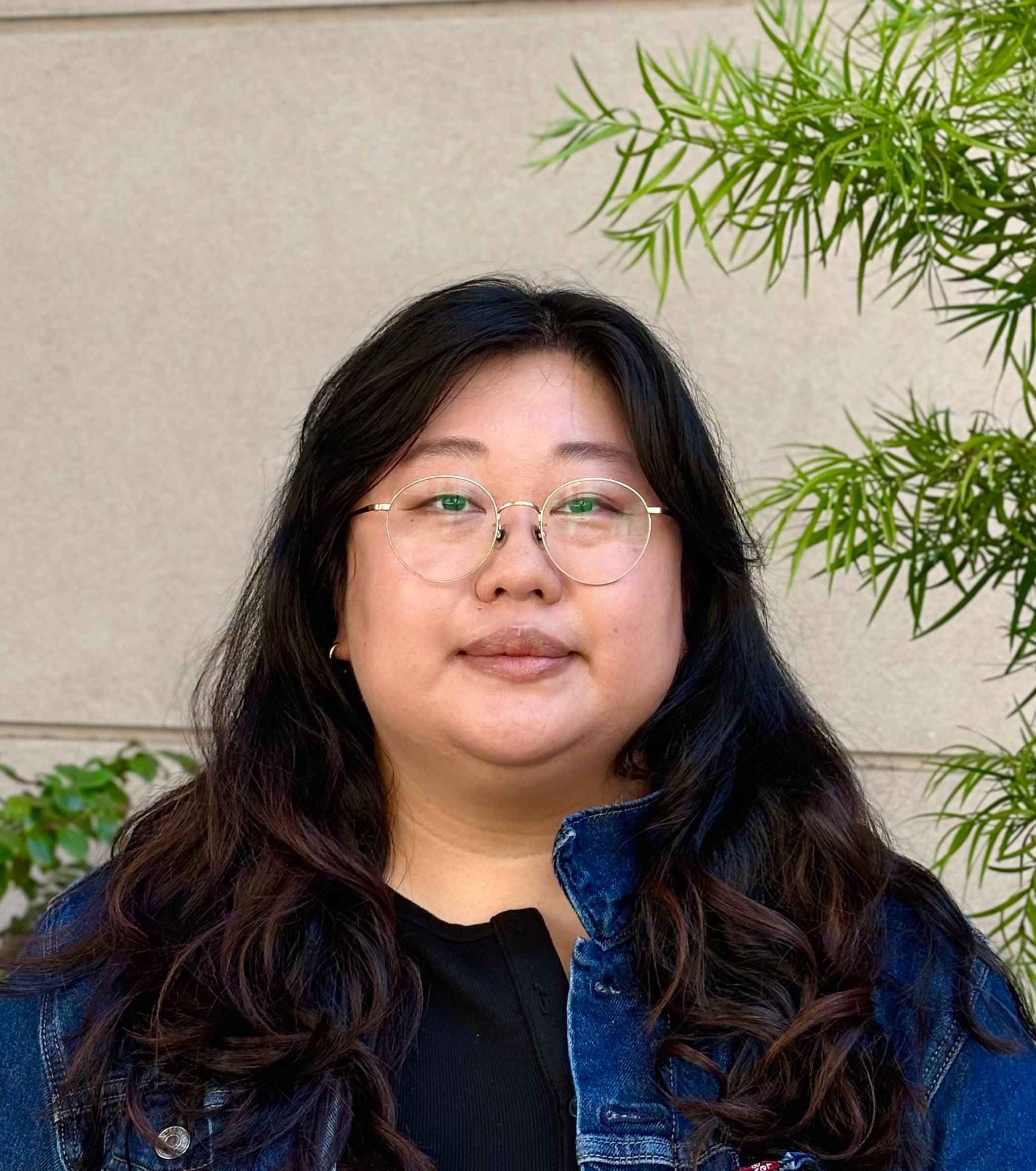 woman with long hair and glasses, wearing a jean jacket, posing in front of a wall with plants in front of it