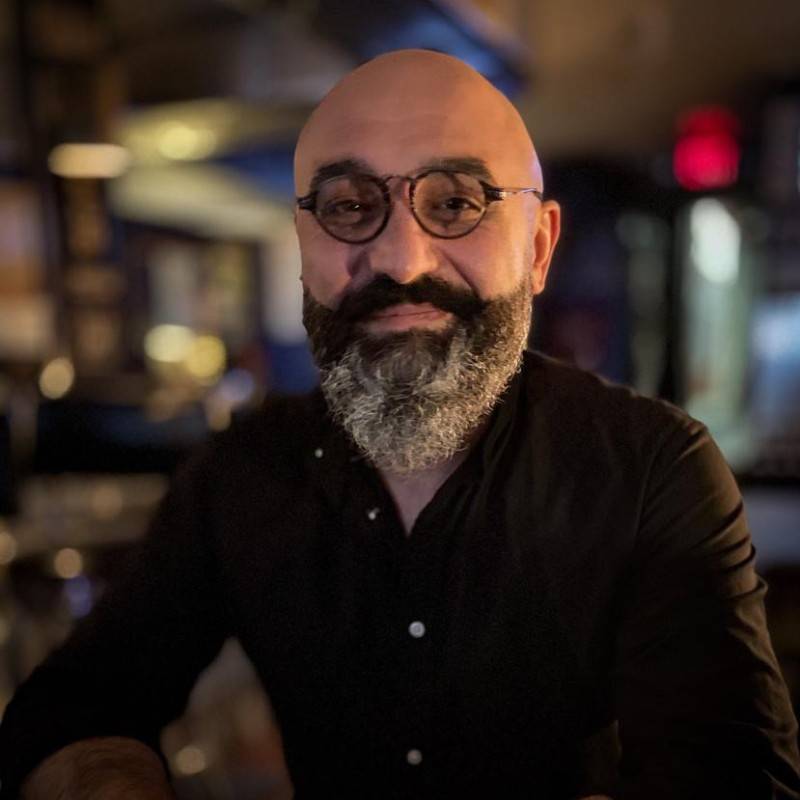 man with glasses, shaved head, salt-and-pepper beard, black shirt, blurred background