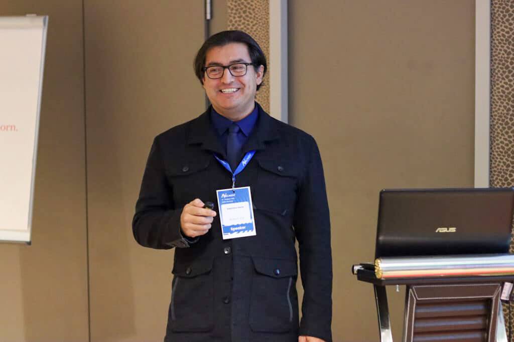 man wearing glasses, dark jacket, blue shirt and tie, standing next to computer, lanyard around neck, looking to the right, smiling