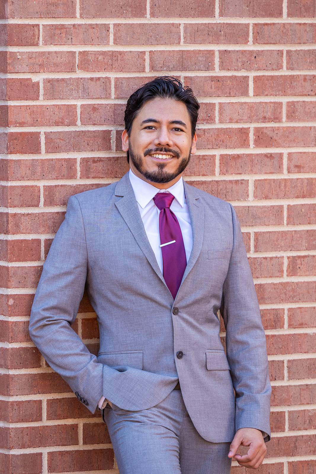 man with beard and mustache, one hand in pocket, grey suit and purple tie, standing in front of brick wall