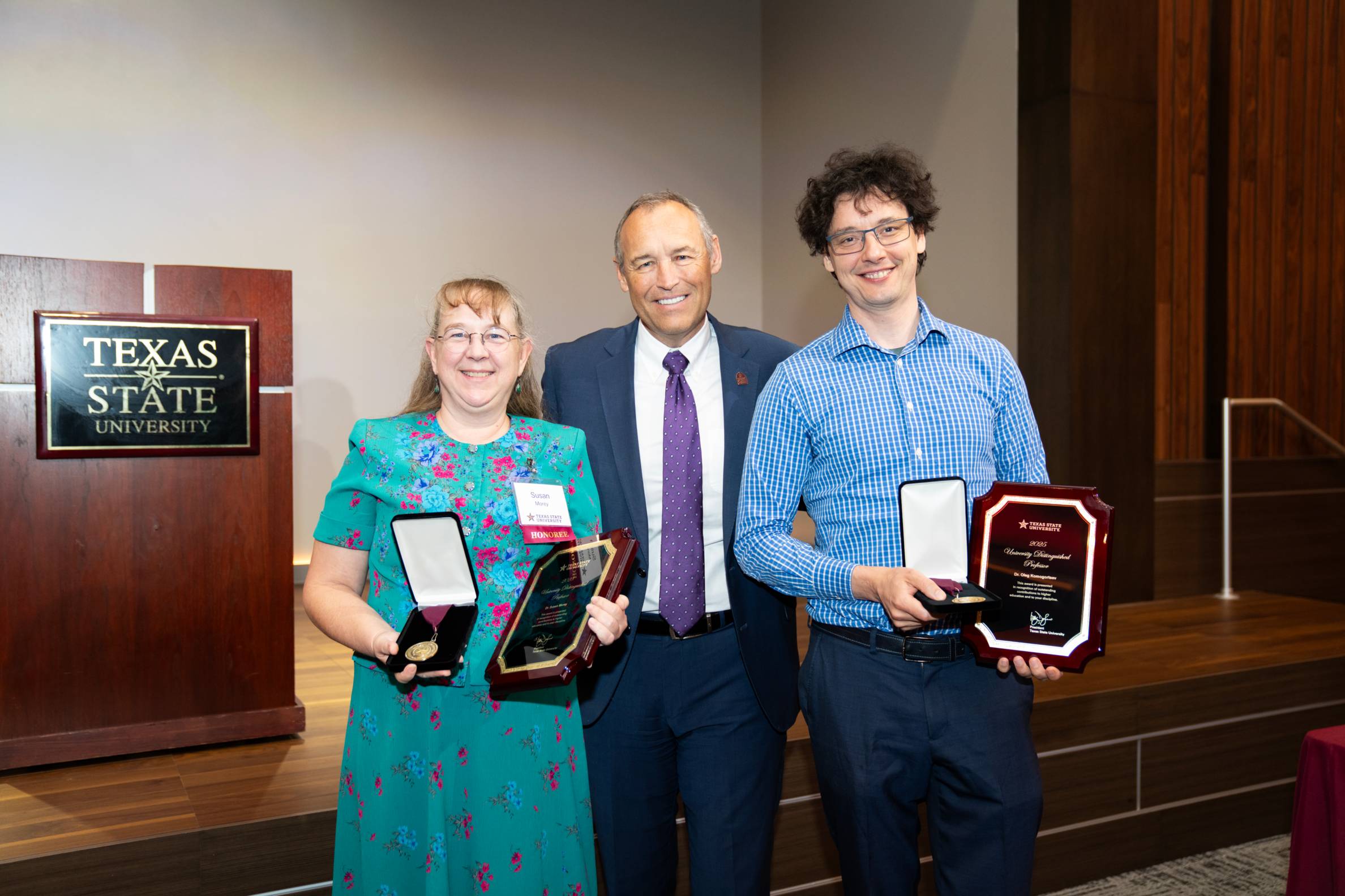 Two faculty award winners hold their plaques while standing on either side of President Kelly Damphousse.