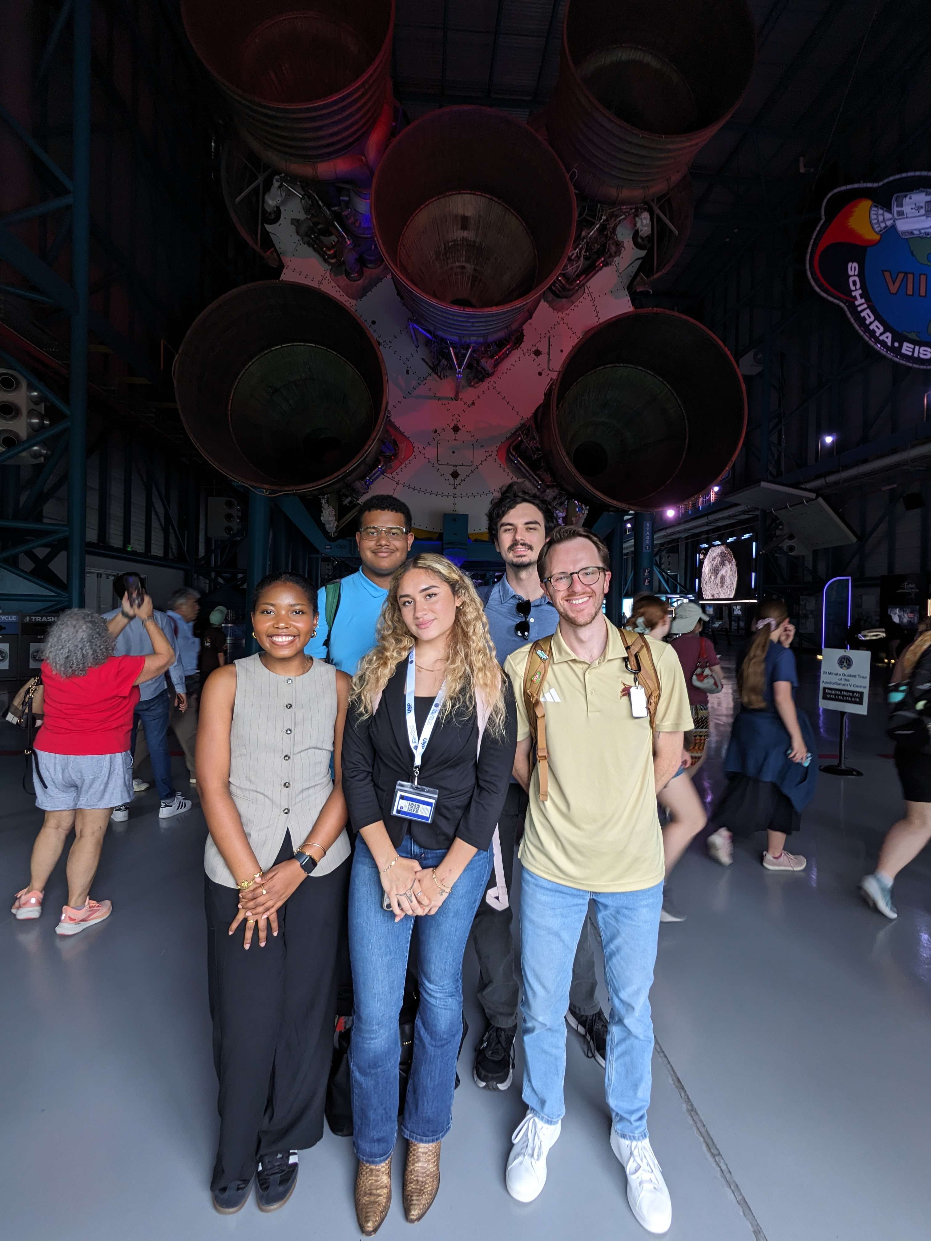 five people standing in front of large rocket in warehouse