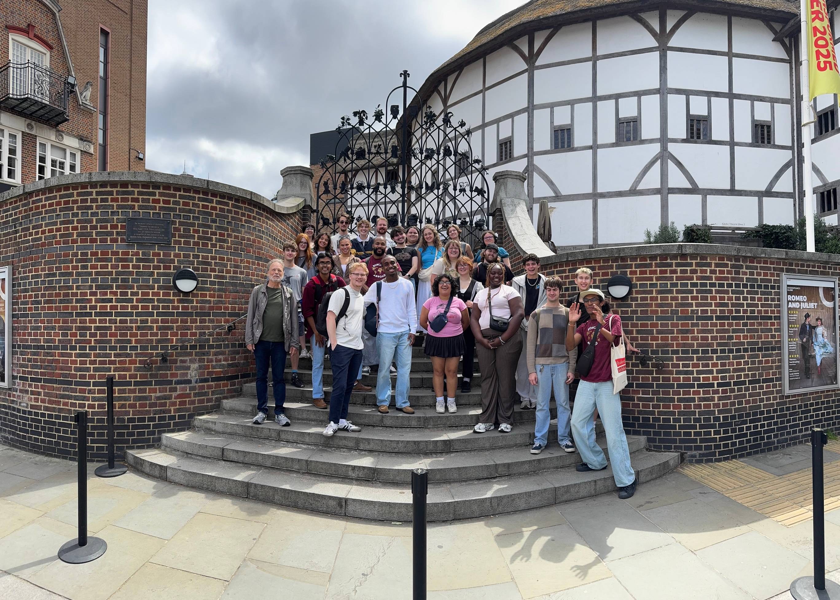 A large study abroad cohort smiling and posing together on stone steps outside the iconic white and timber-framed Shakespeare's Globe theater in London.