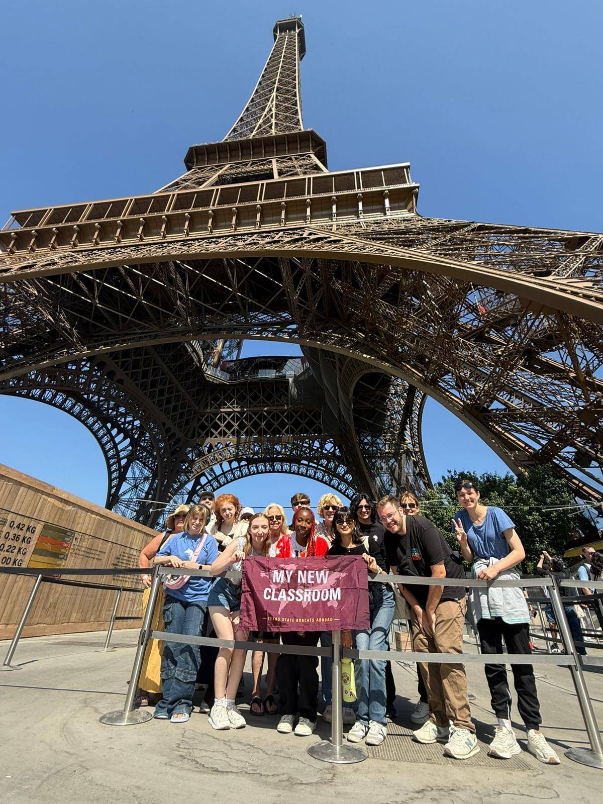 A large group of students and instructors smiling and posing for a photo directly underneath the massive iron legs of the Eiffel Tower.
