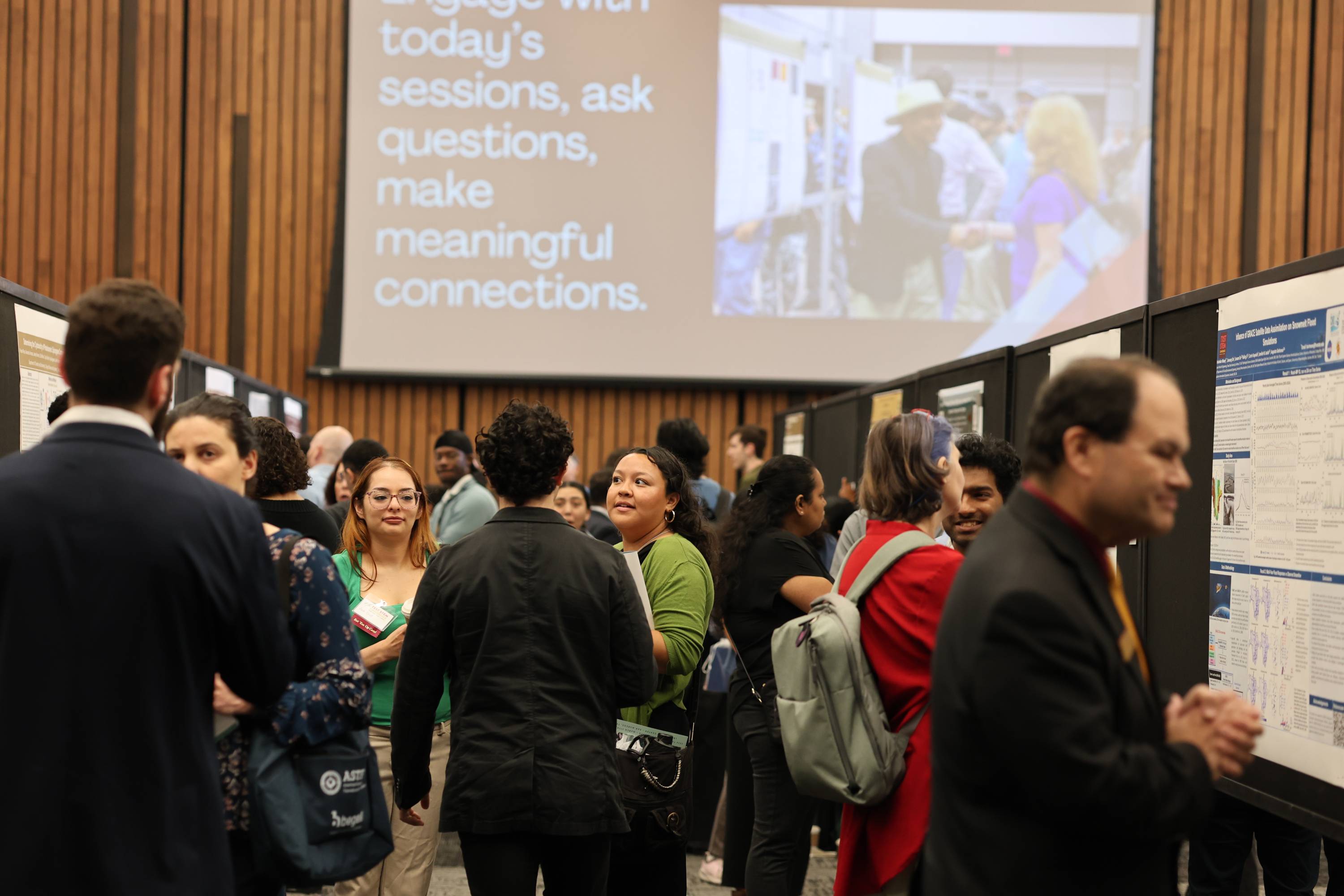Attendees mingle among research poster boards at an academic conference, with a projector screen overhead displaying the text "Engage with today's sessions, ask questions, make meaningful connections." alongside a photo of people networking at a similar poster session.