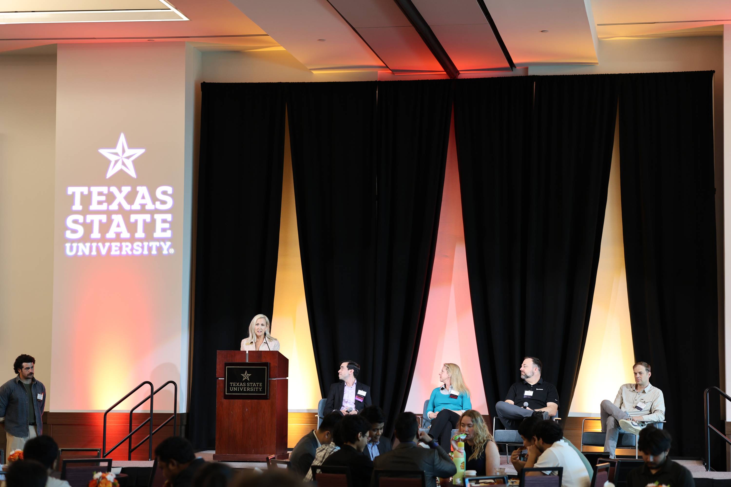 A woman speaks at a wooden podium bearing the Texas State University seal, with four panelists seated beside her on stage and the university's star logo projected on the wall behind them, while an audience watches from below.