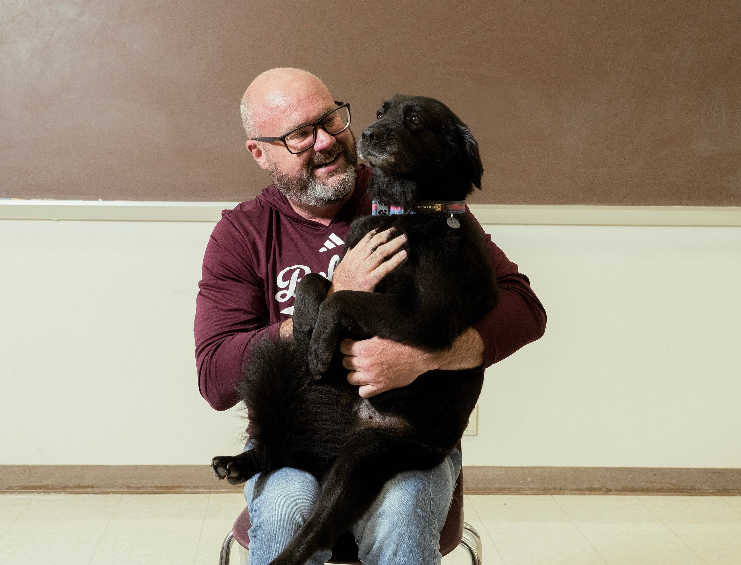 Dale Blasingame with his dog Lucy sitting in his lap like a big baby. Dale is wearing a maroon sweatshirt with Bobcats written on it. Lucy is a medium sized dog with black fur and a little bit of gray on her muzzle.