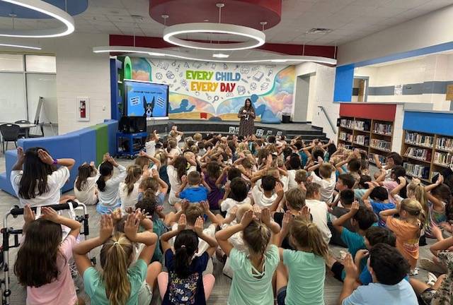 woman reading a book to a large group of children who have their hands up on their heads to mimic dog ears
