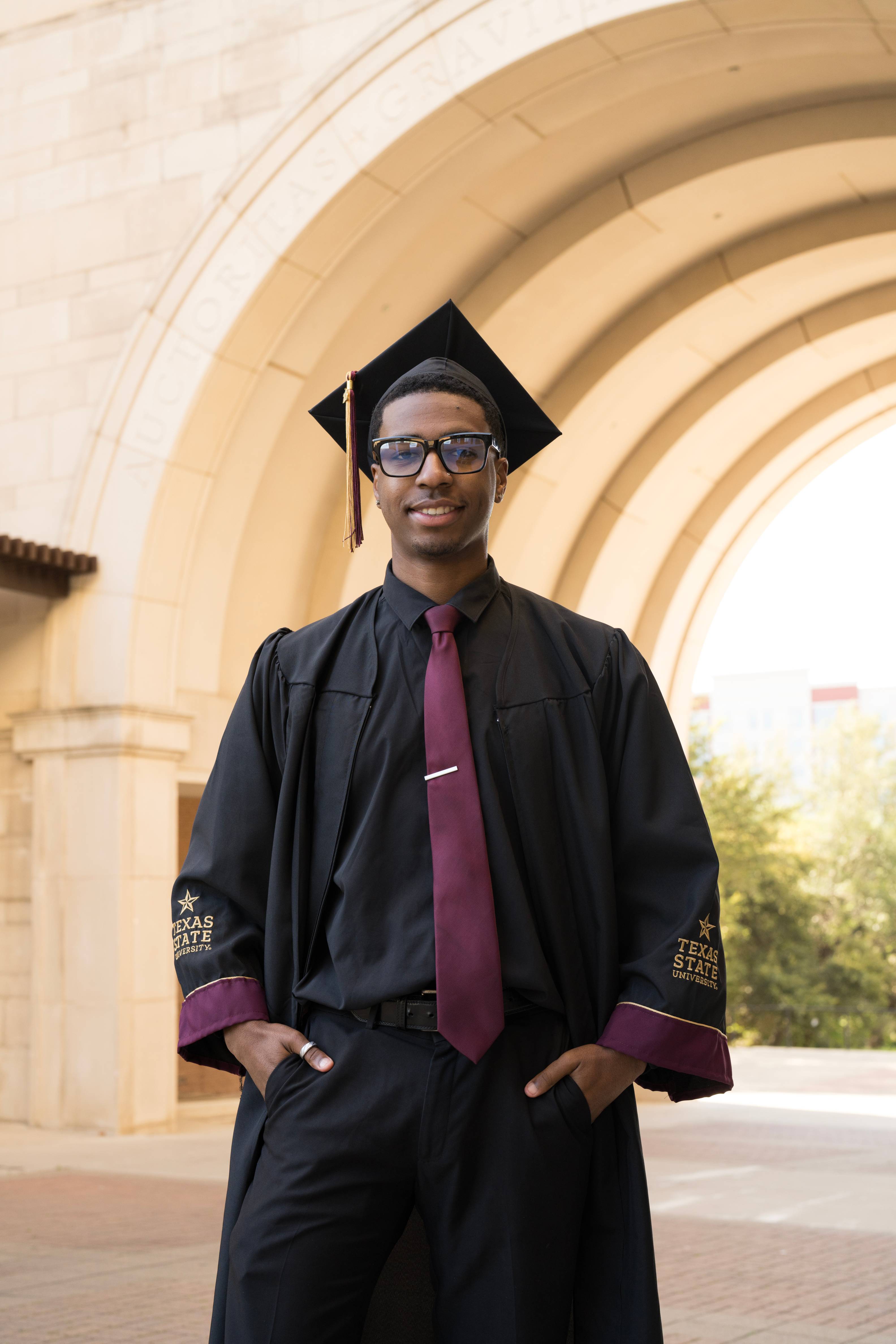 kaison wearing glasses and regalia smiles at the camera with the archway in the background