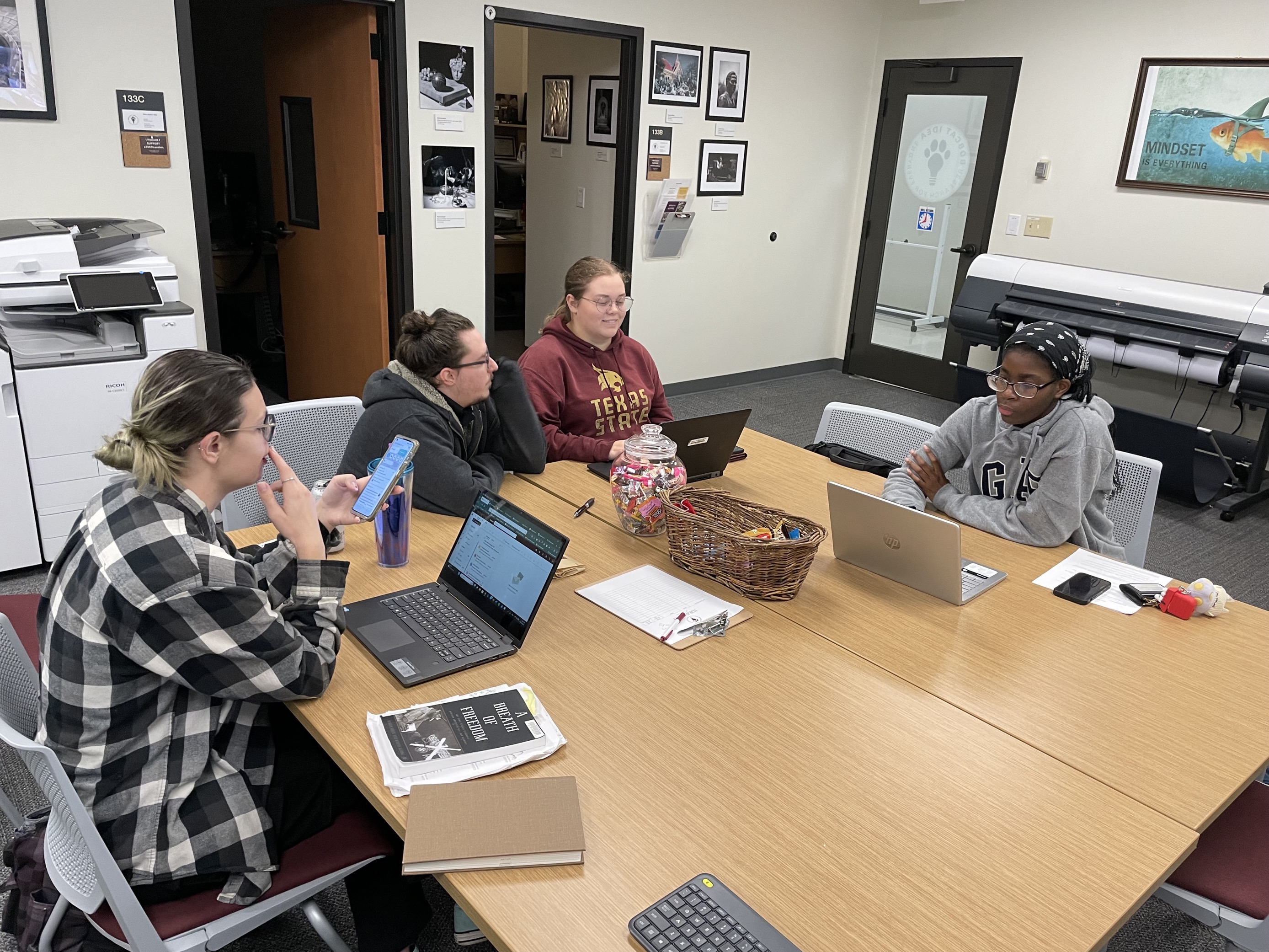 staff member hosting a meeting. three students are sitting around a table with laptops while discussing their work with a staff member. 