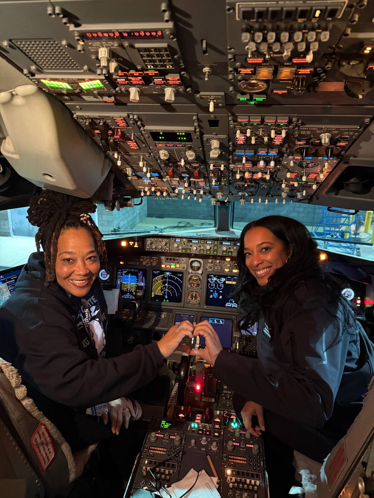  Two women in the cockpit of an airplane. They are joining their hands together to make a heart shape.