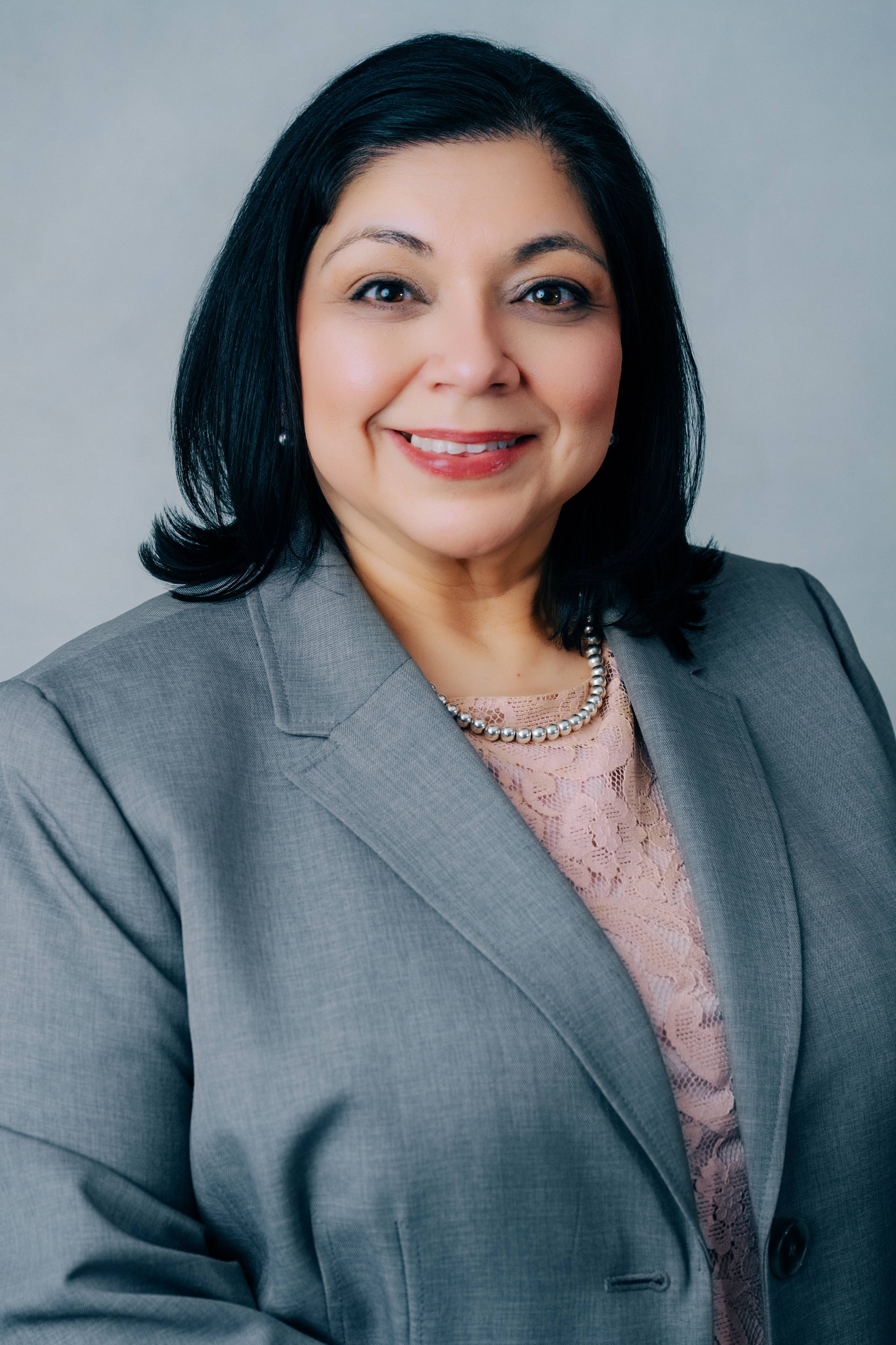 Professional headshot of a smiling woman with shoulder-length black hair wearing a light gray blazer over a pink lace blouse and a pearl necklace. She faces the camera against a soft gray background, conveying a polished, approachable, and confident appearance suitable for a business profile, speaker bio, or leadership page.