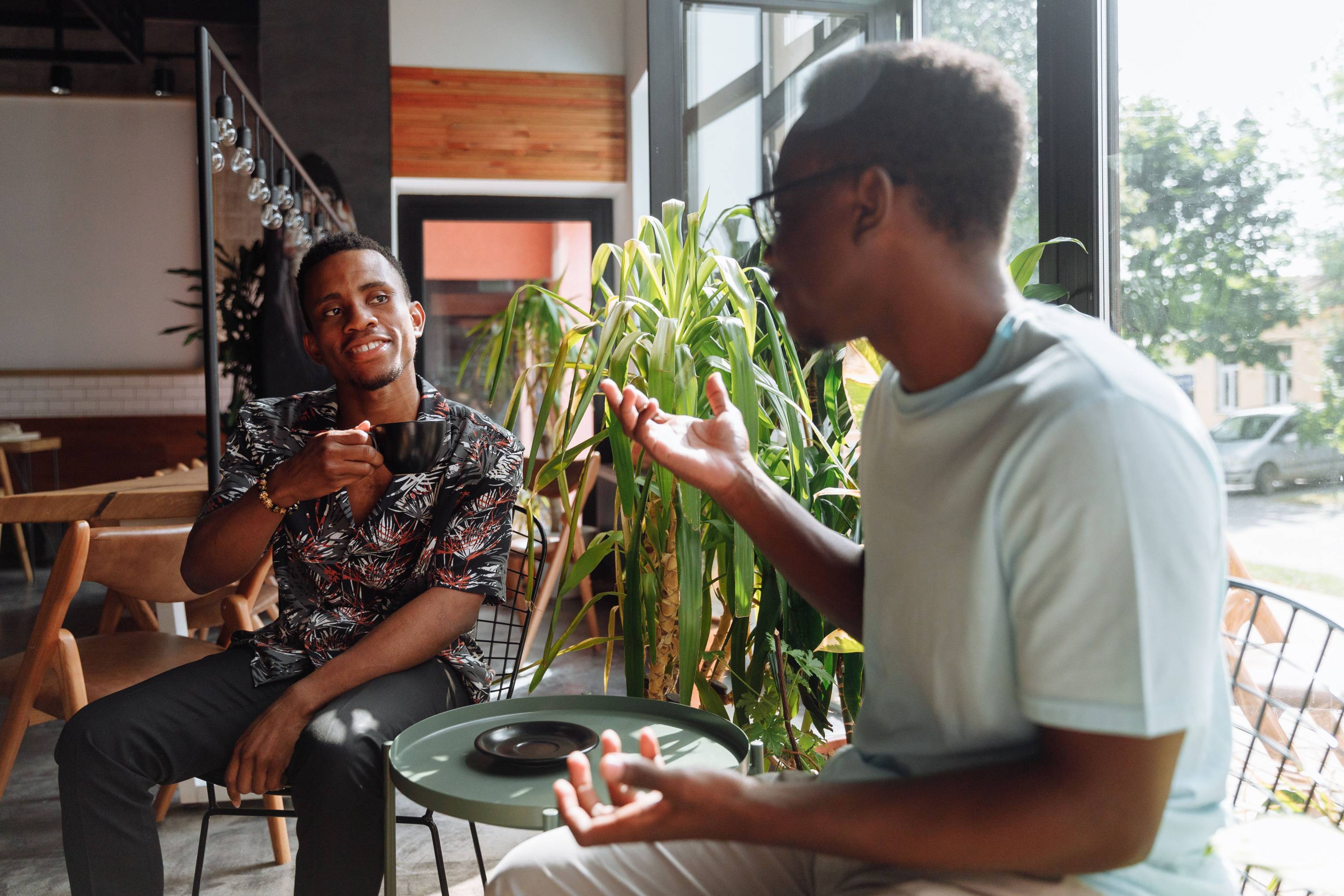 Two people chat over coffee at a café, smiling and engaged in conversation. One gestures while the other listens, creating a friendly, relaxed atmosphere. The image represents networking, relationship building, customer referrals, word-of-mouth marketing, and growing a business through personal connections and trust.