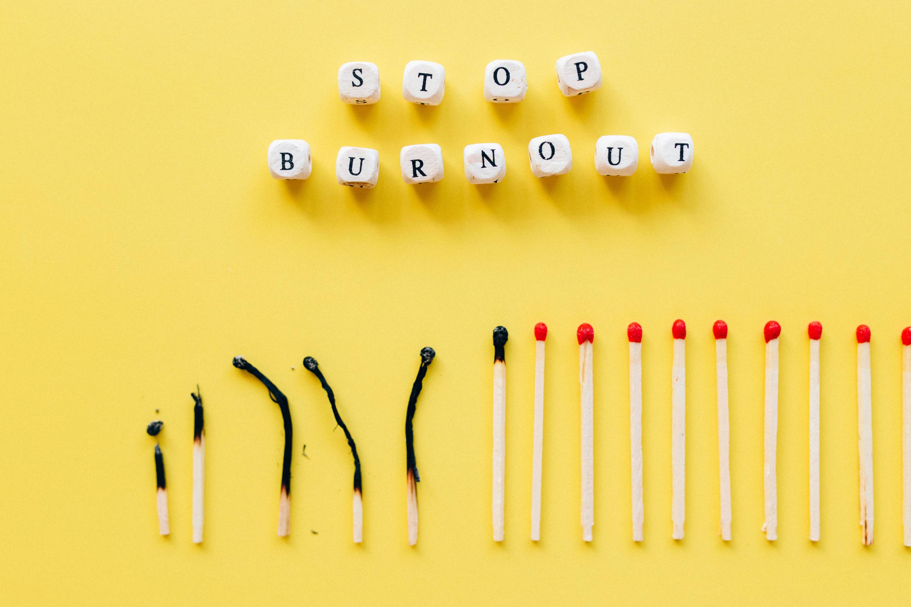Graphic with the words "Stop Burnout" spelled using wooden letter blocks on a bright yellow background. Below, a row of matchsticks transitions from burned and charred to unlit, symbolizing recovery, resilience, workplace wellness, stress management, mental health, and preventing professional burnout.