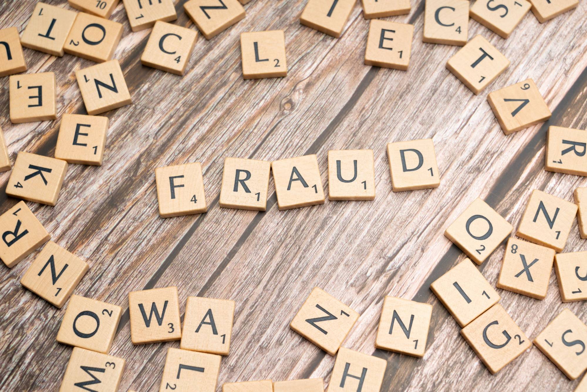 Wooden letter tiles on a rustic wood surface spell the word “FRAUD,” surrounded by scattered alphabet tiles. The image symbolizes fraud prevention, financial scams, identity theft, cybersecurity awareness, risk management, and protecting businesses and consumers from fraudulent activity.