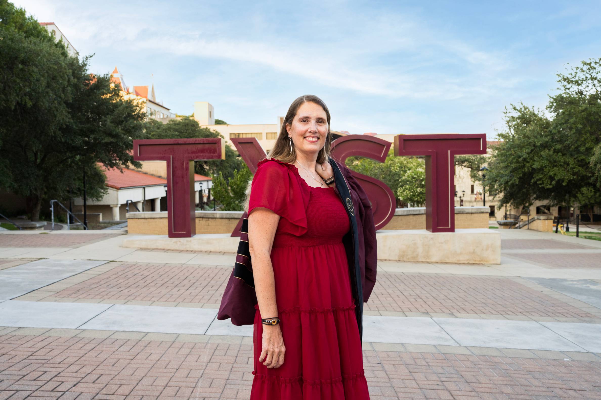 beth wears a bright red dress and poses in front of the txst letters sign