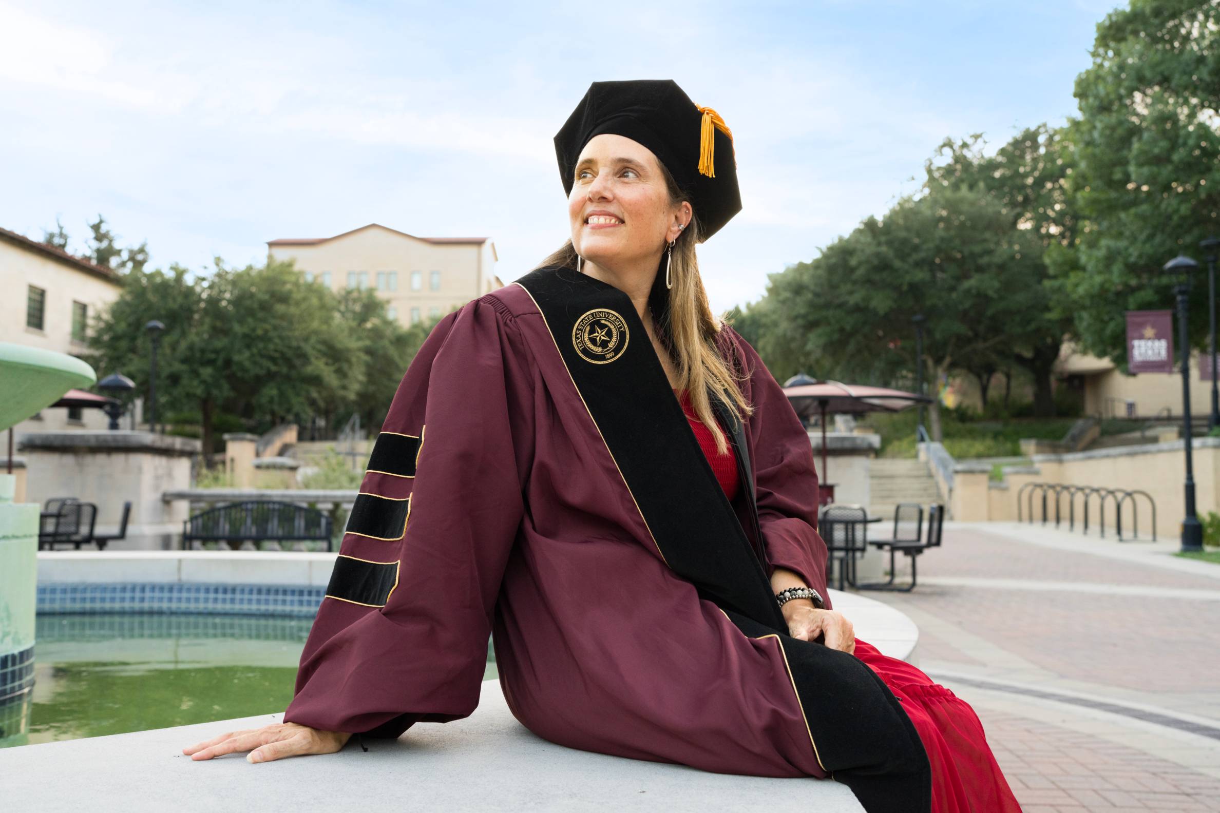 beth wearing graduation cap and gown perches on the edge of a fountain and looks into the distance