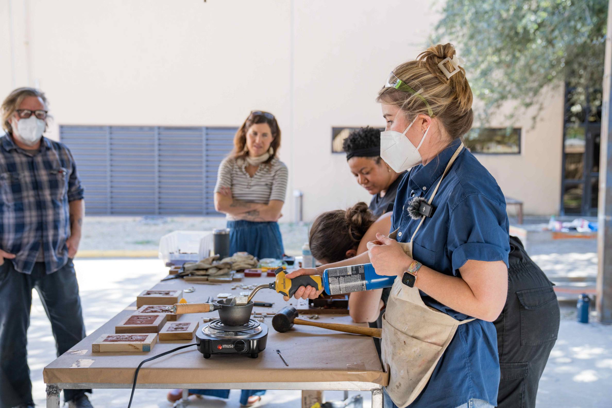 woman with mask and apron works over table of equipment outside while bystanders look on