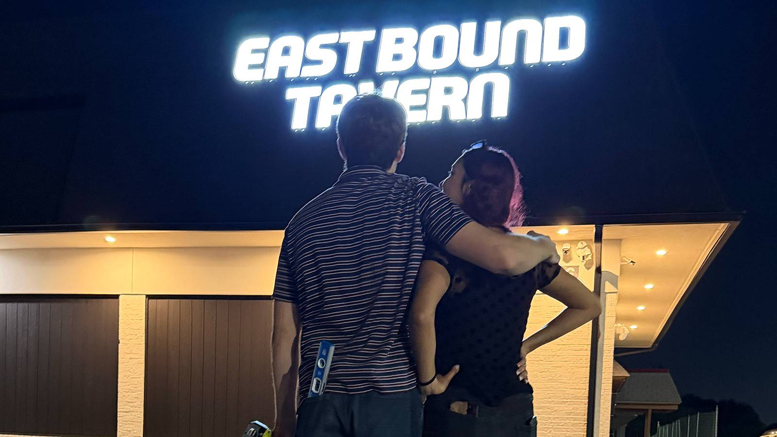 man hugs a woman over her shoulders as they stare at a building that reads Eastbound Tavern