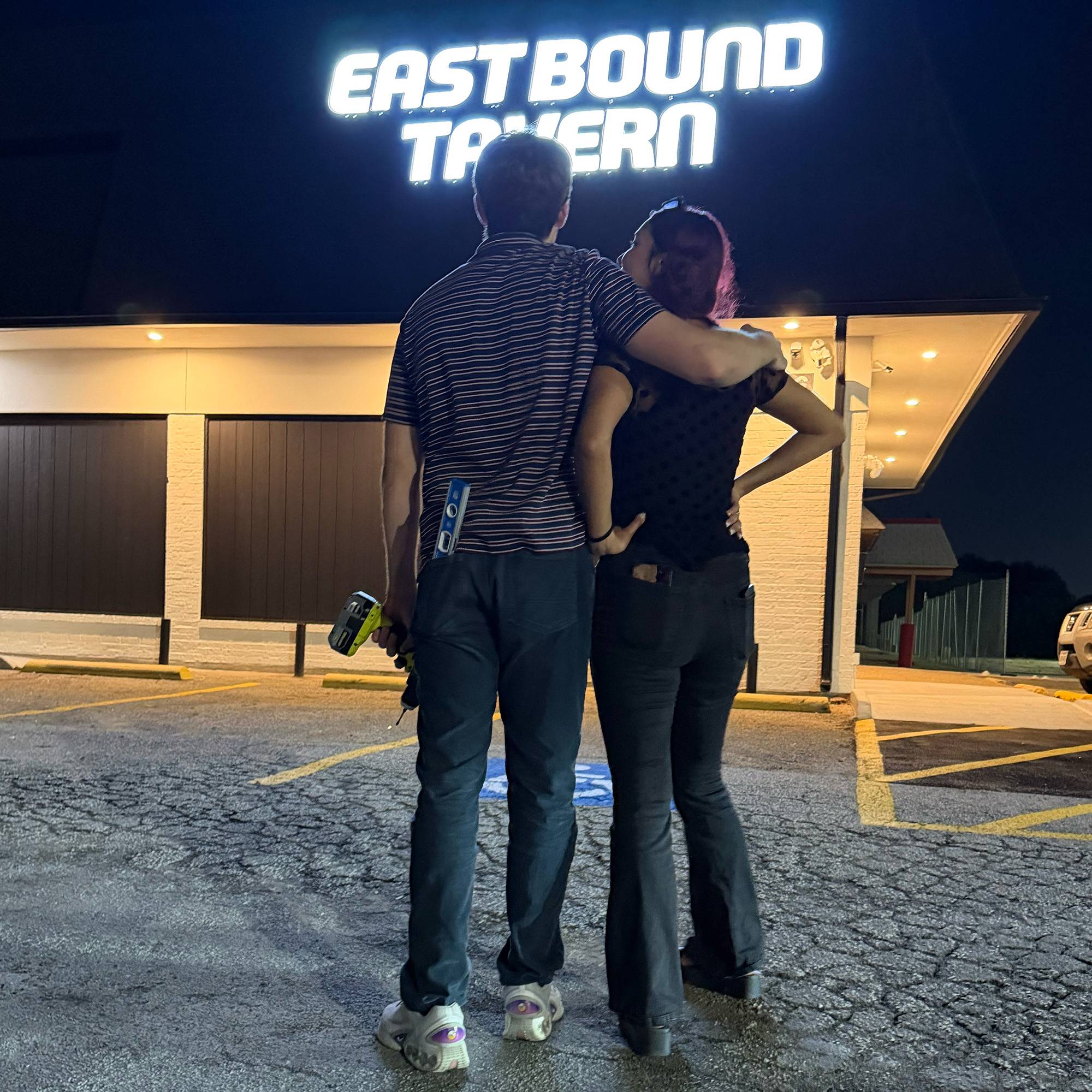 man hugs a woman over her shoulders as they stare at a building that reads Eastbound Tavern