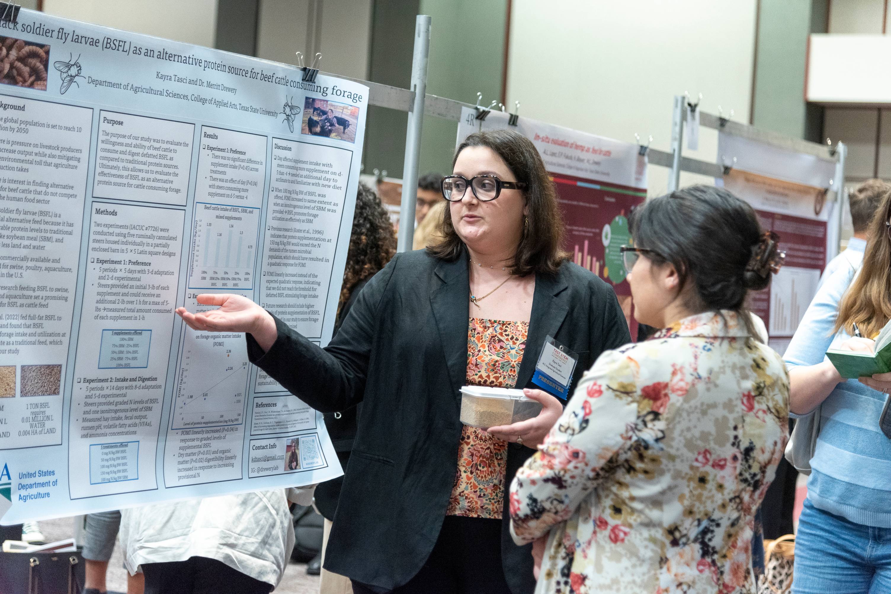  a woman in a blazer pointing to her research poster, speaking to another woman