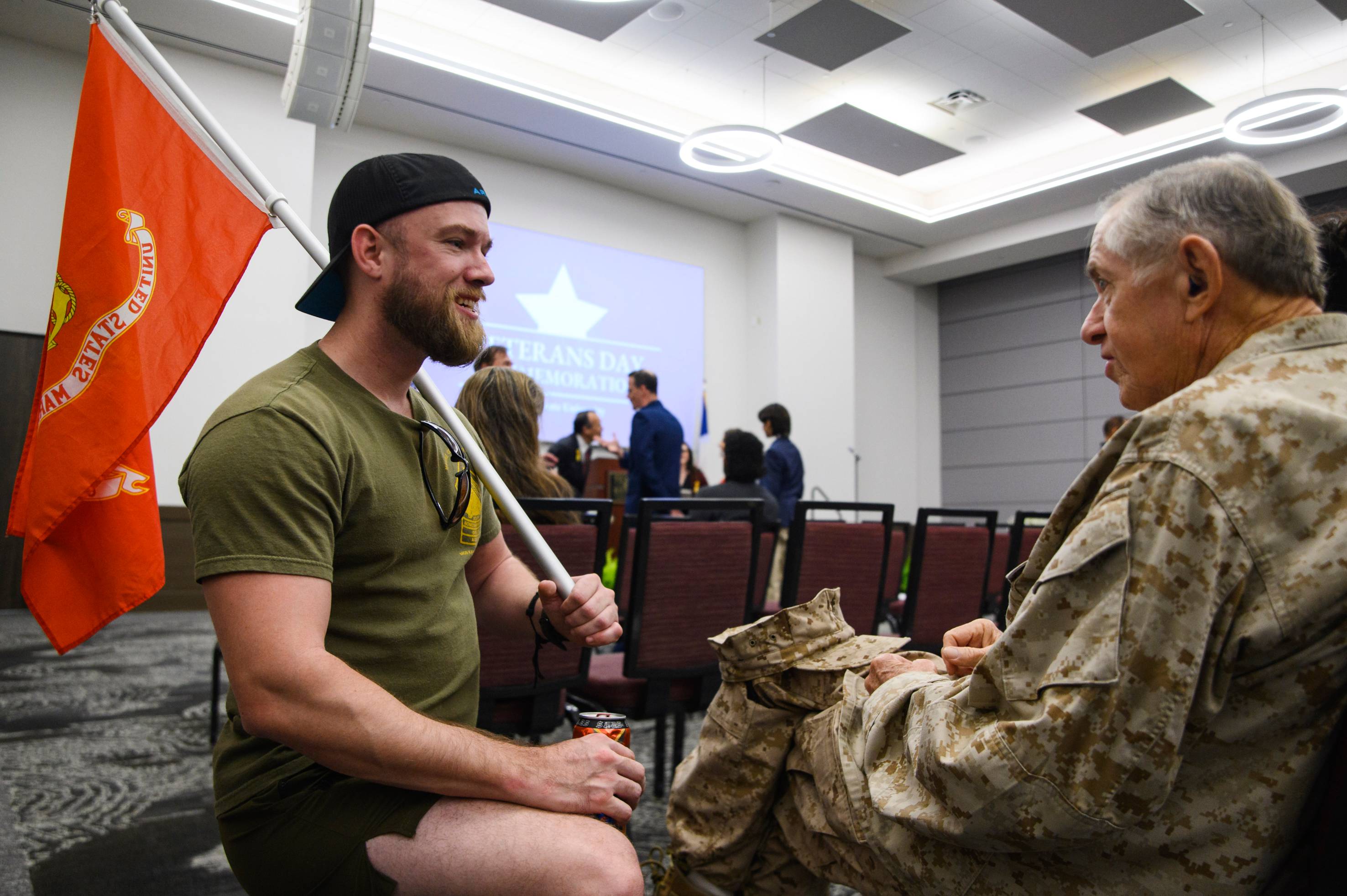 young man holding a marine corps flag kneeling next to older man sitting while in uniform 