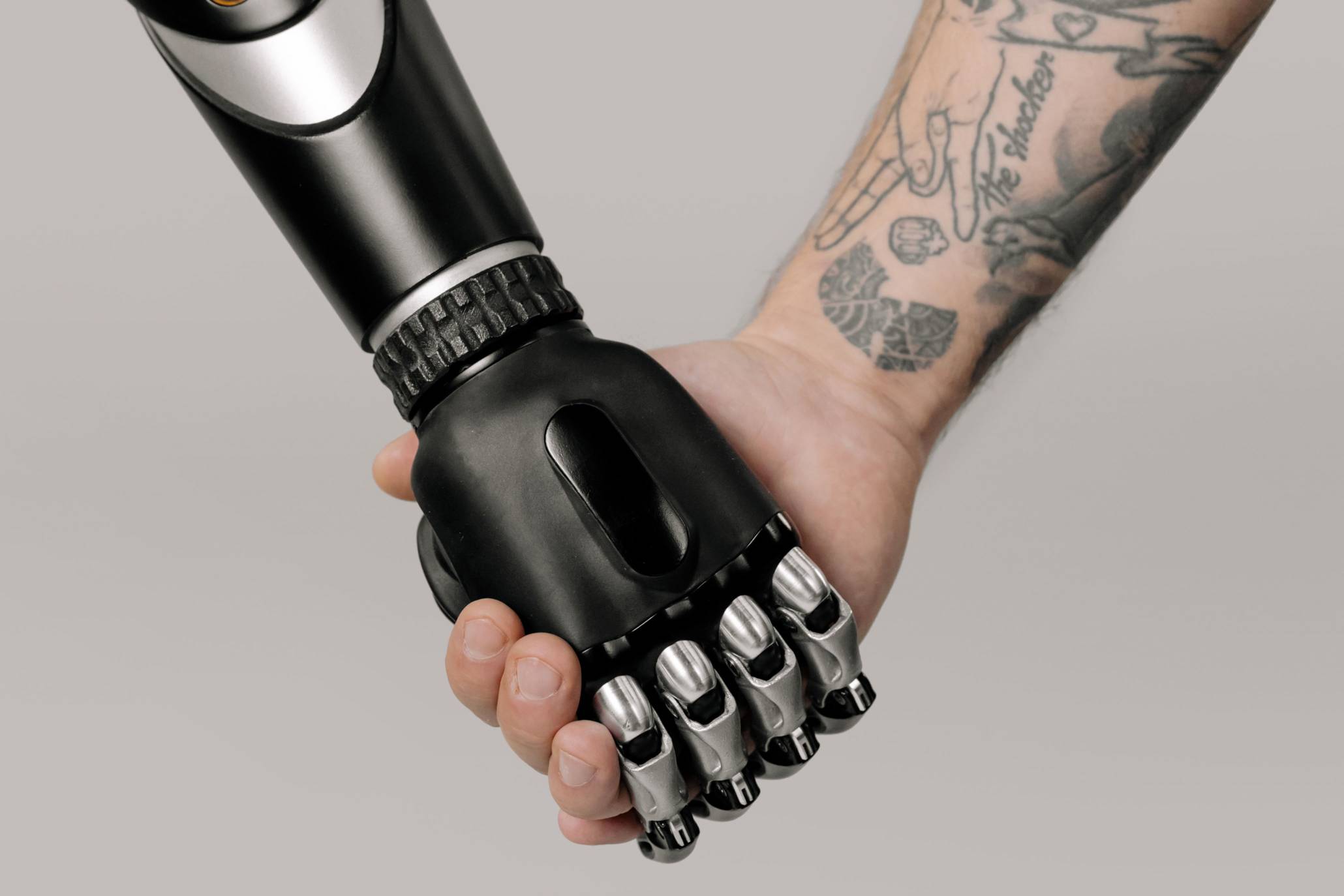 Close-up of a person shaking hands with a robotic prosthetic hand against a neutral background. The image symbolizes human-technology collaboration, artificial intelligence, assistive technology, innovation, accessibility, and the growing role of advanced technology in work and everyday life.