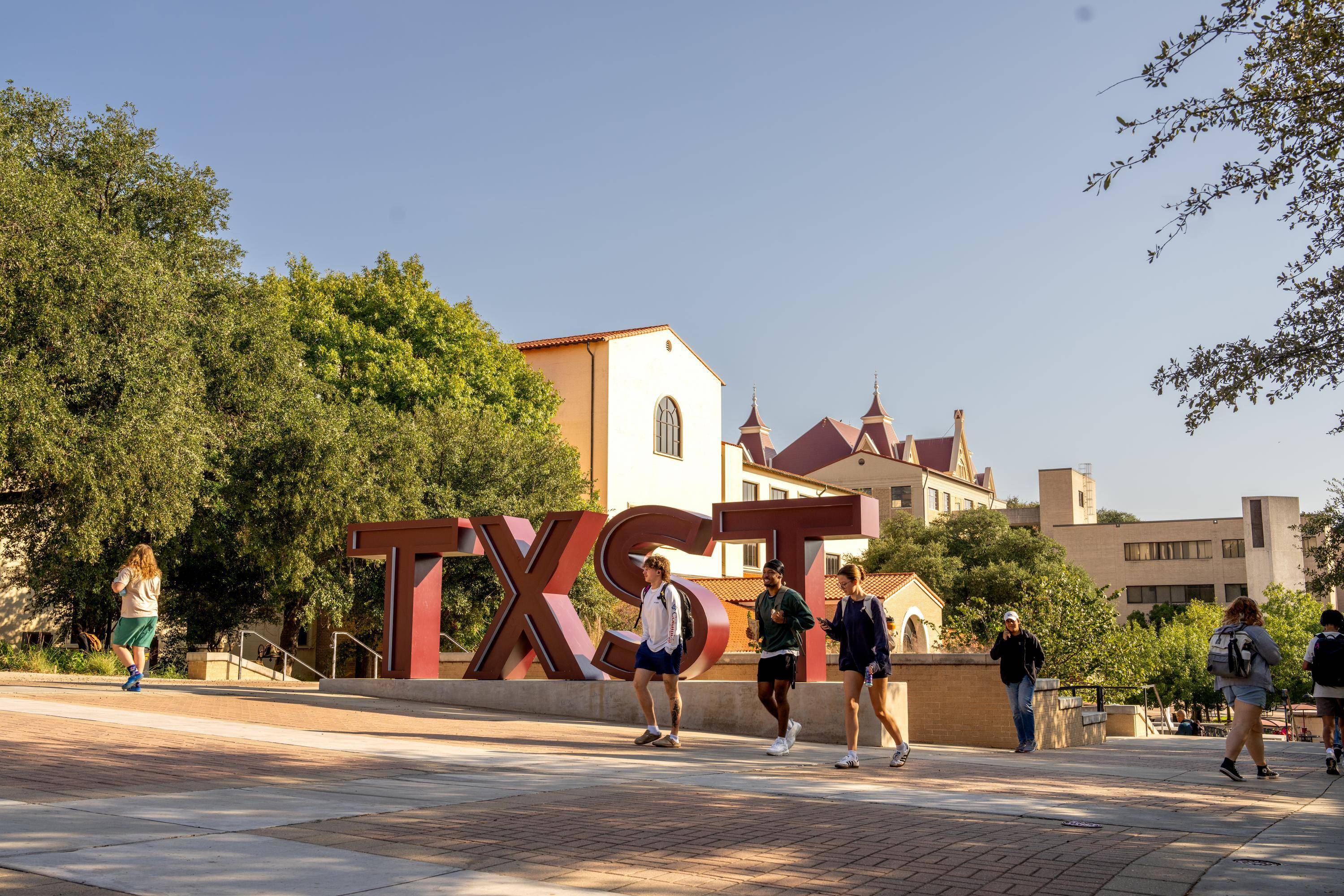 students walking on campus in front of large maroon TXST sign