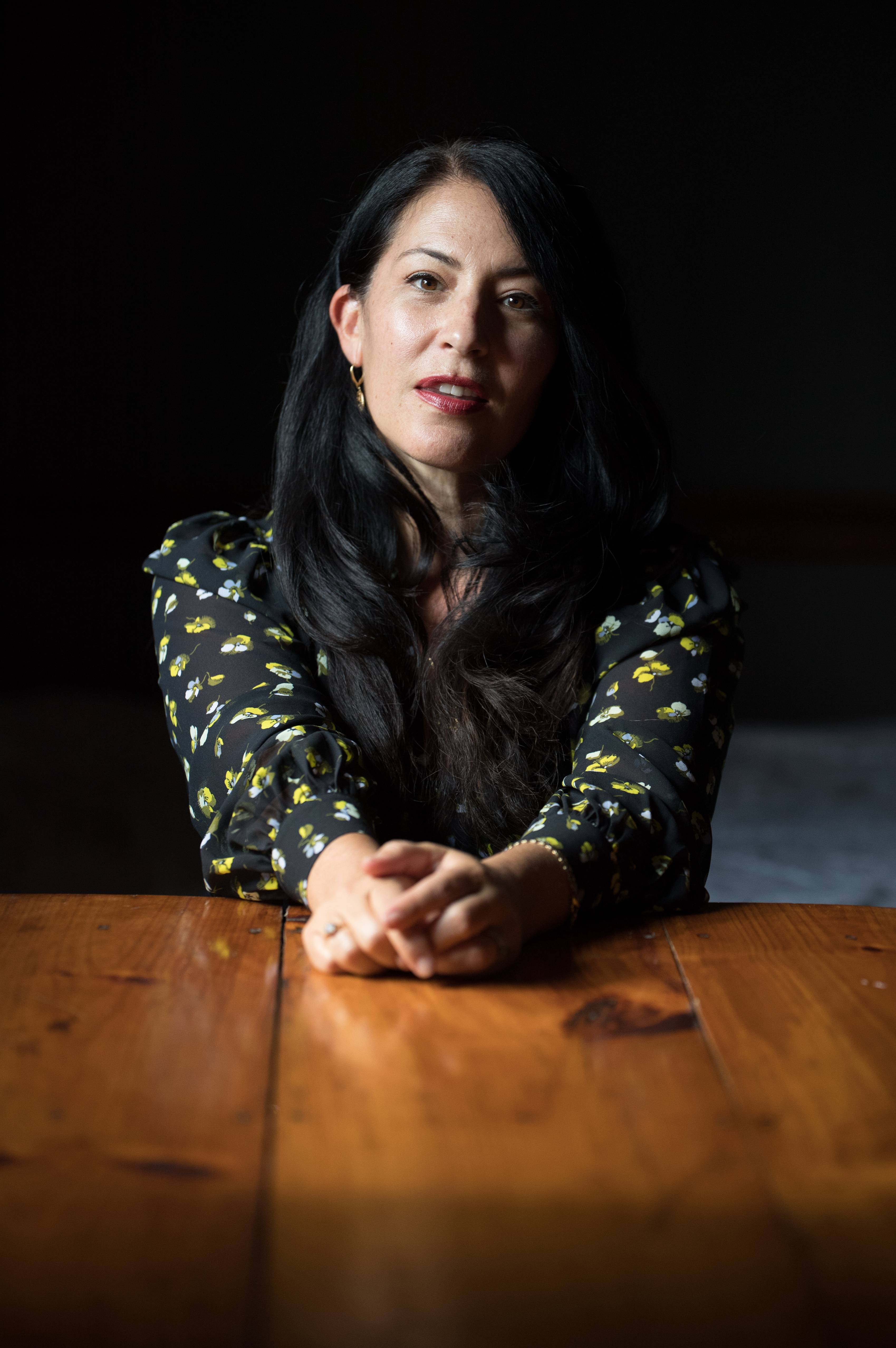 Ada Limon sitting at wooden desk with her arms out on the desk. the lighting is dark and dramatic