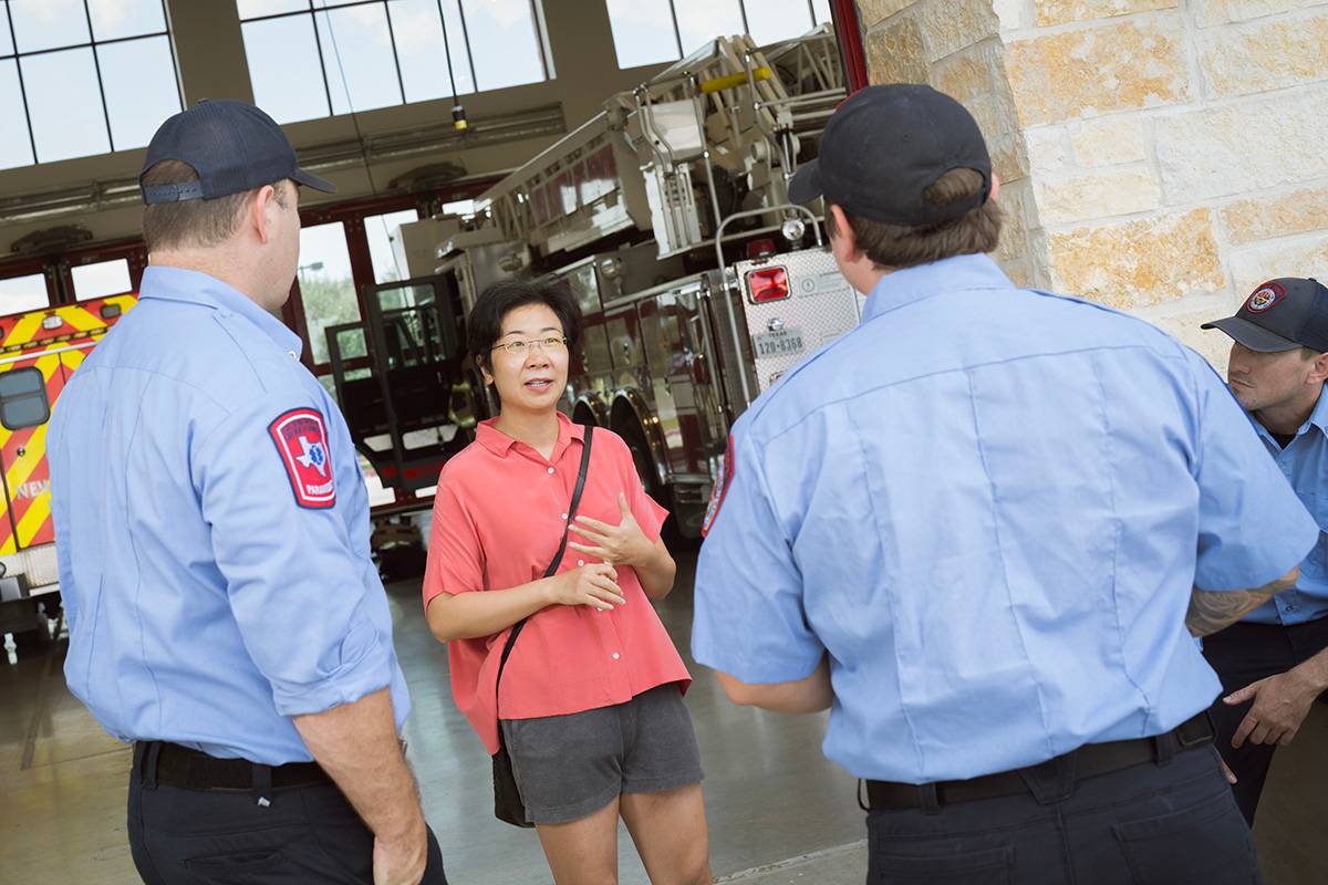 Dr. Xi Pan standing, talks to a group of fire fighters in front of a garage with fire trucks in them.