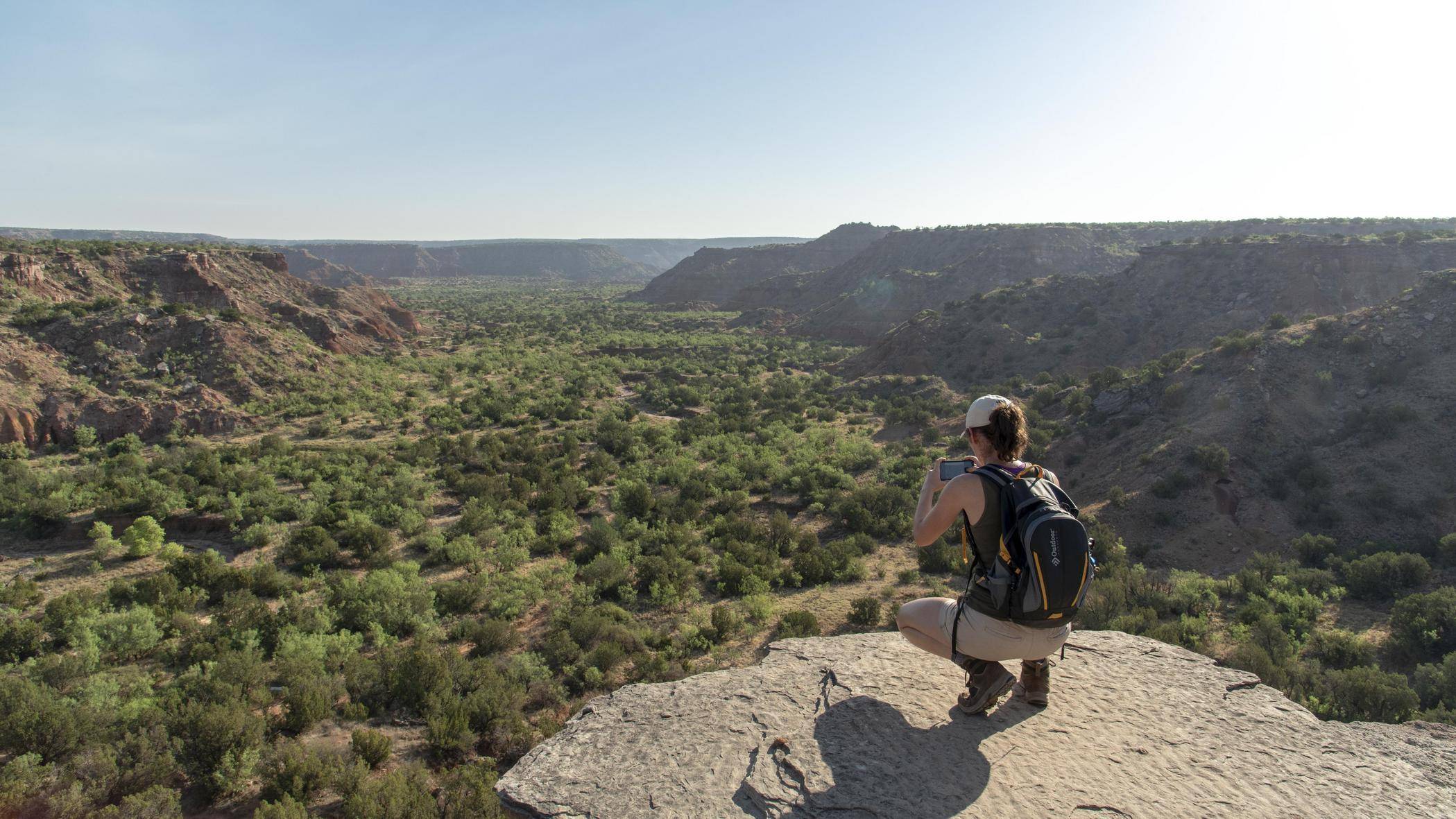 Young woman in hiking gear crouches to snap a photo of Texas Hill Country.
